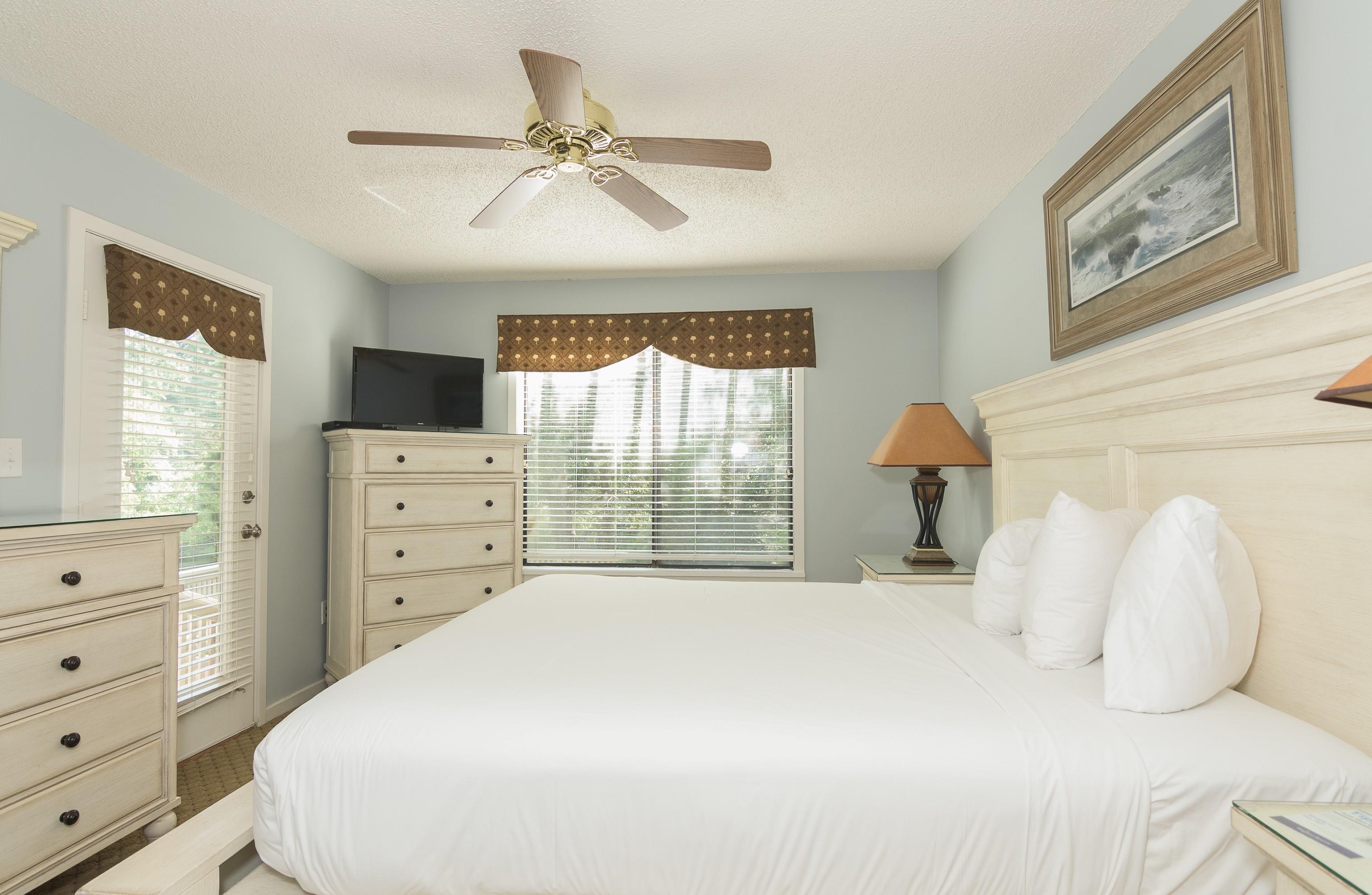 Furnished bedroom with a white bed, pale wood furniture, and windows with patterned valances.