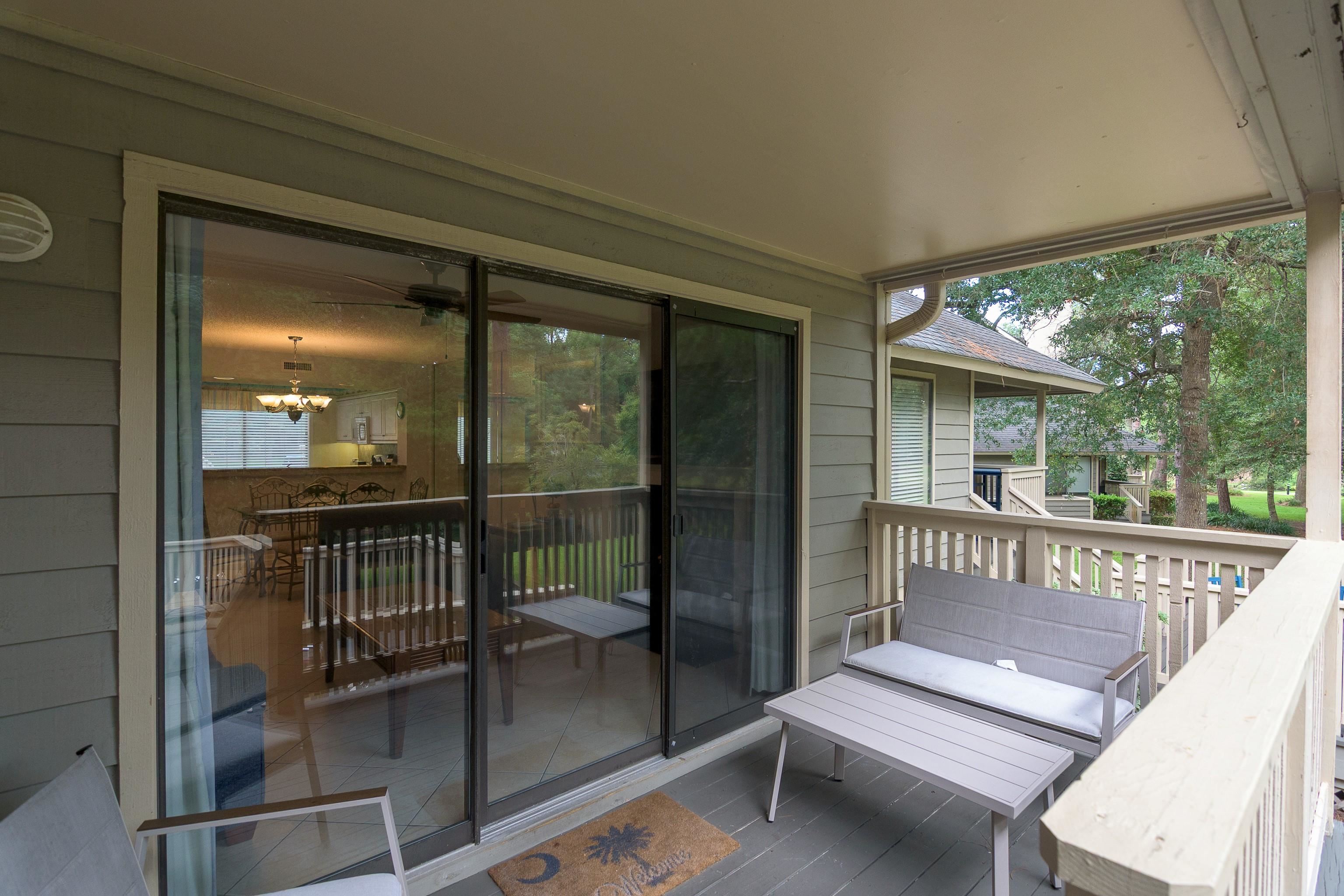 Covered balcony with gray lounge seating, sliding glass doors, and views of trees and neighboring homes.