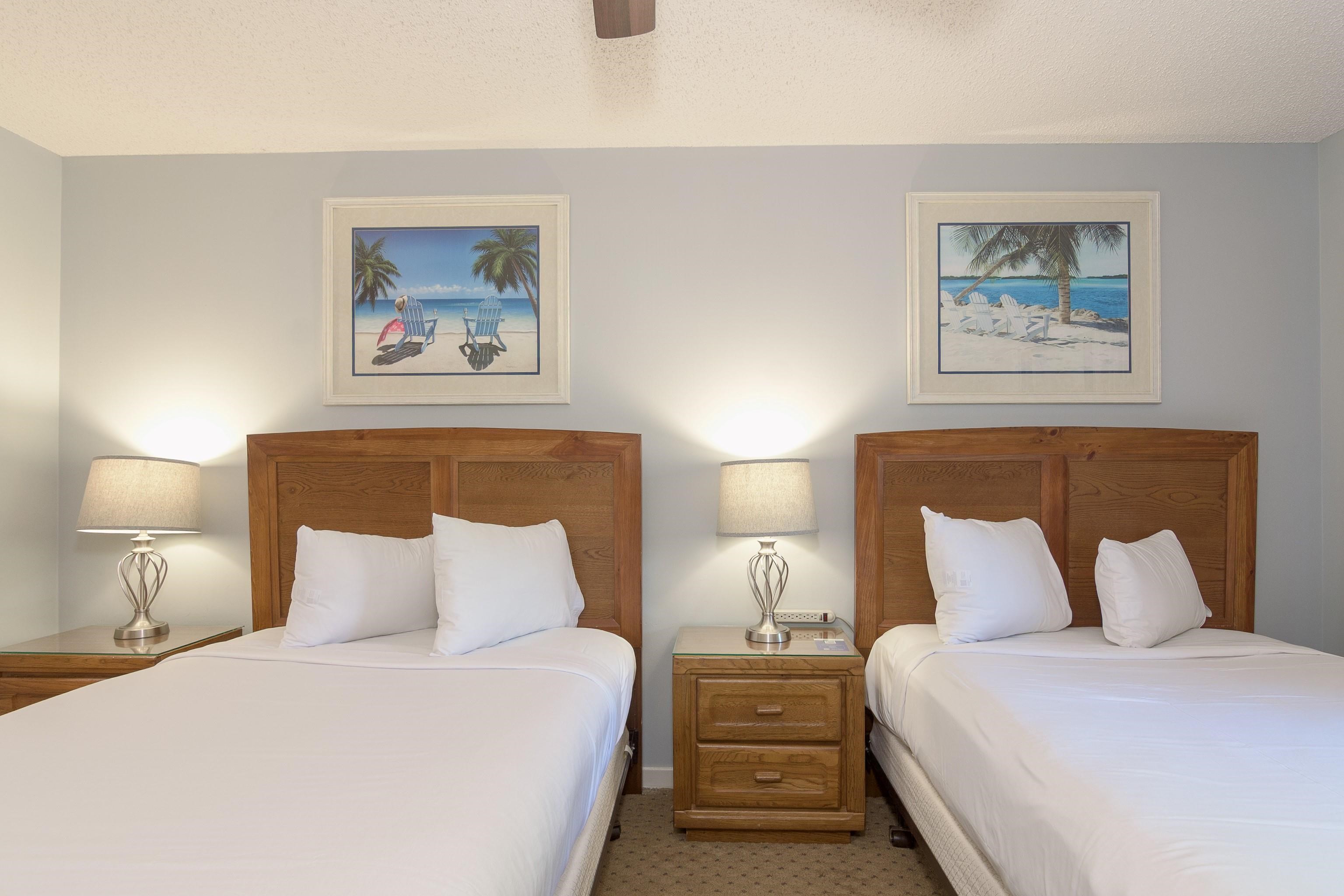 Bedroom with two white beds, wooden headboards, bedside lamps, and framed beach scenes.