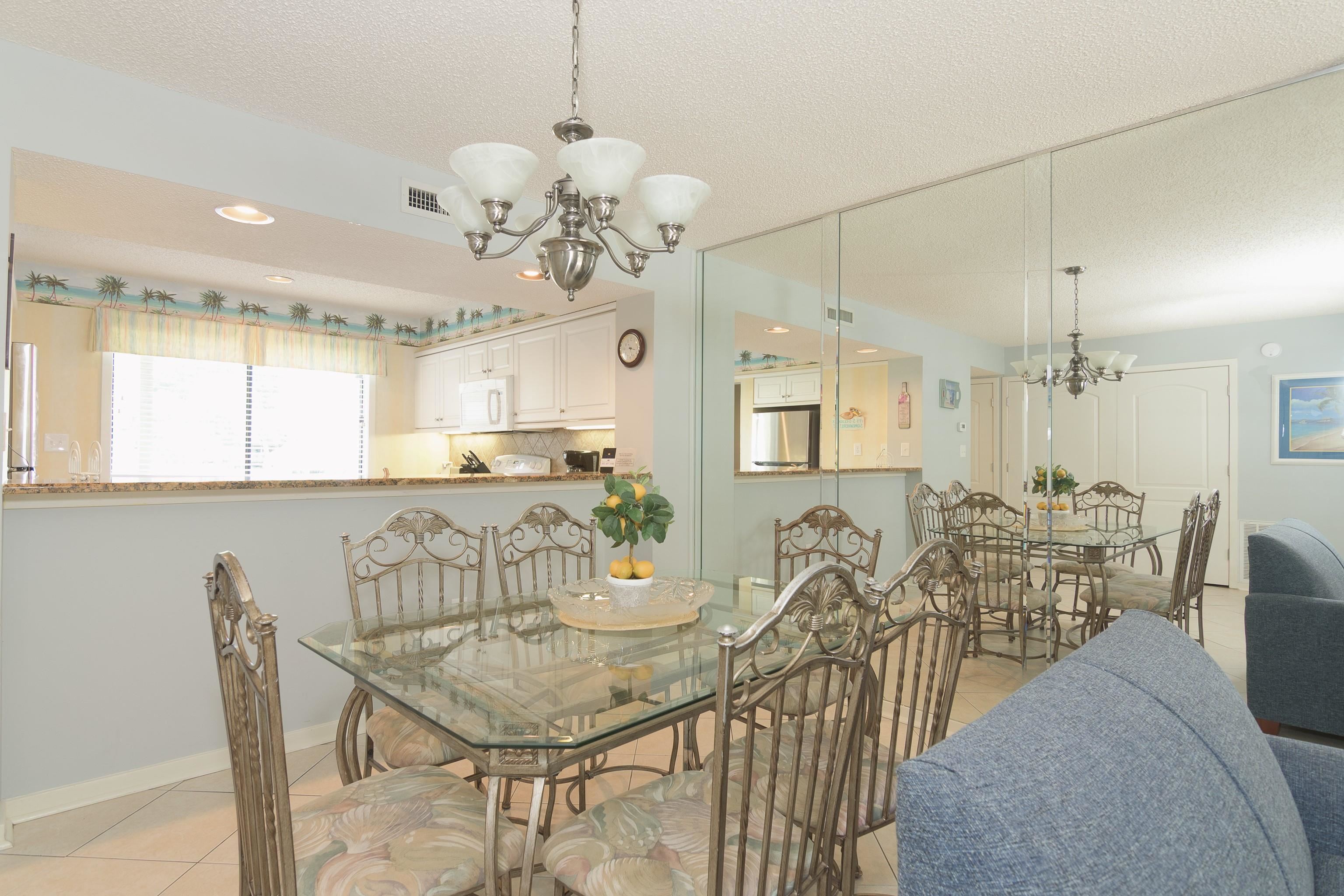Dining area with a glass table, ornate metal chairs, mirrored wall, and kitchen pass-through.