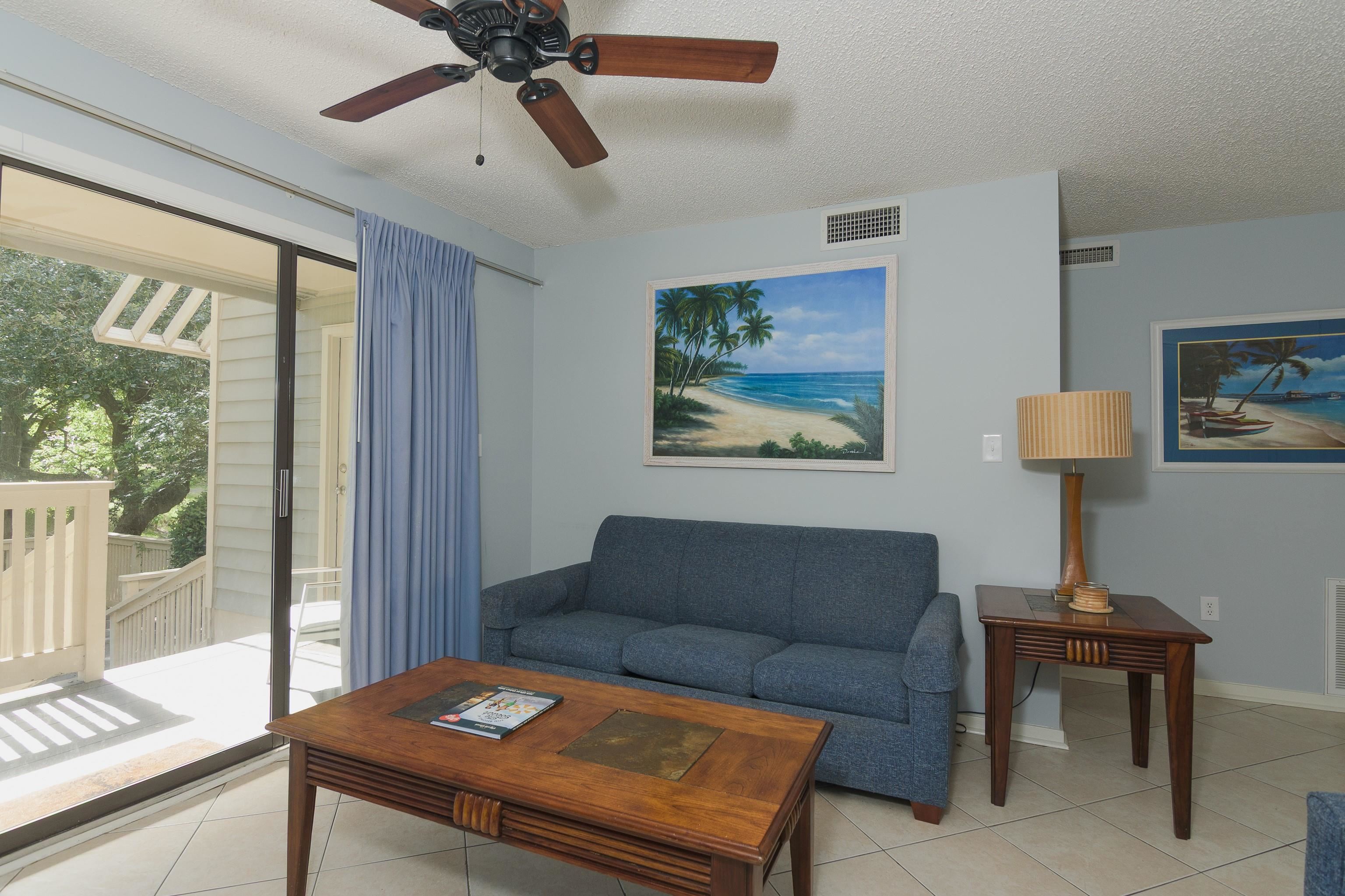 Living room with a blue sofa, wooden furniture, ceiling fan, and sliding doors to a balcony.