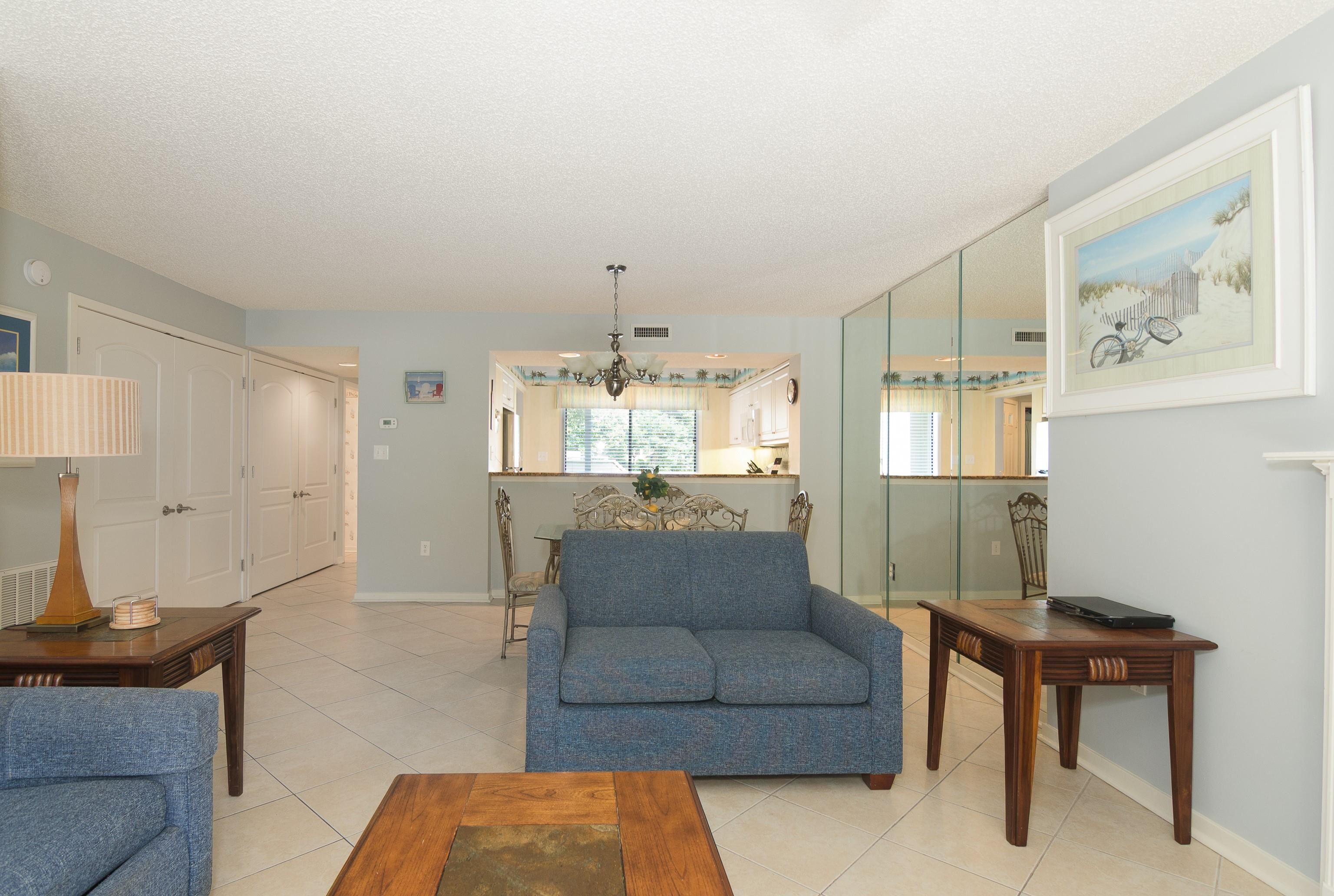 Living room with blue sofas and wood tables, opening to a dining area and kitchen.