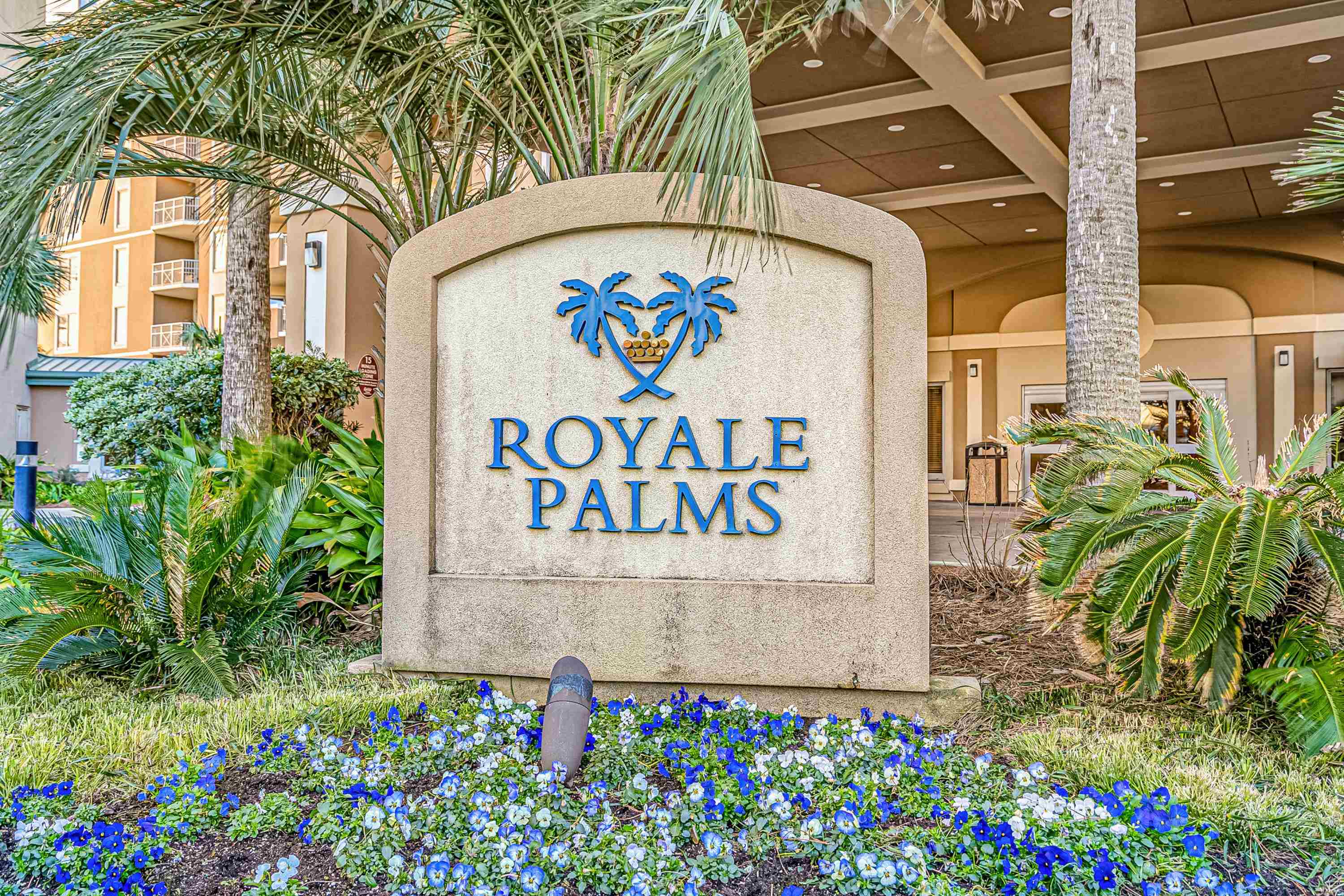 Royale Palms entrance sign surrounded by palm trees and blue and white flowers.