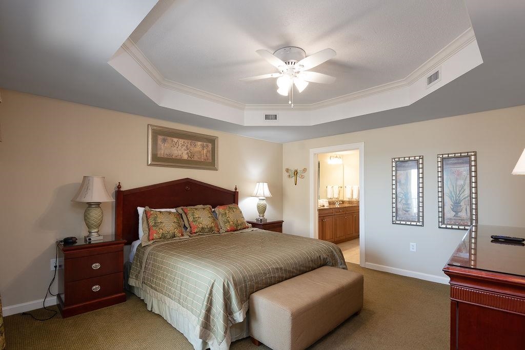 Furnished bedroom with a large bed, tray ceiling, ceiling fan, and open doorway to a bathroom.