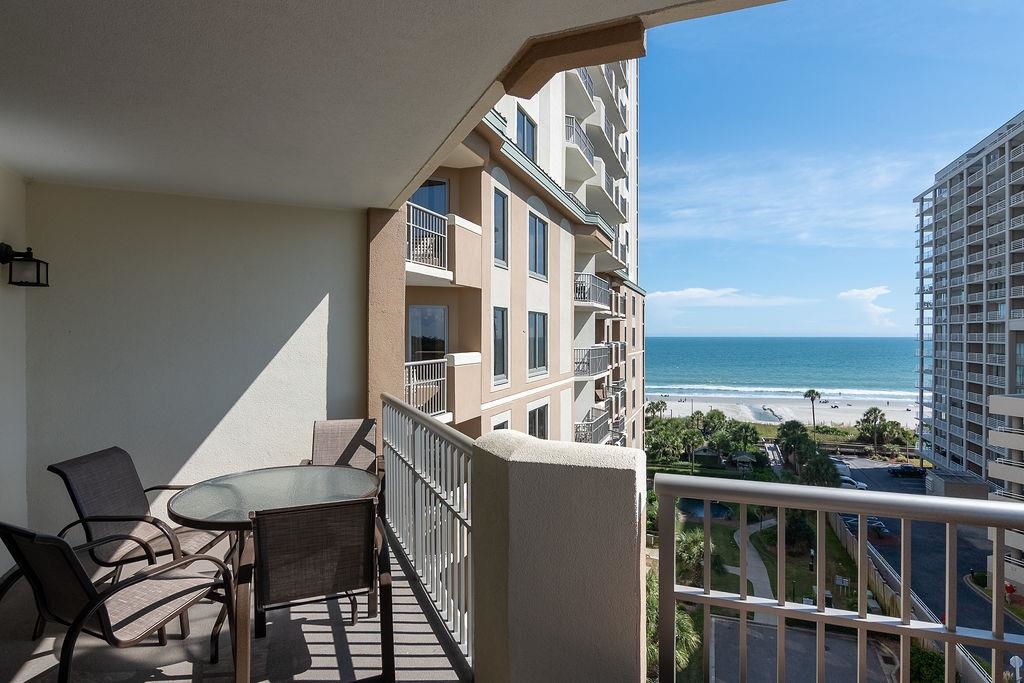 Furnished condo balcony overlooking the beach and ocean, with a glass table and chairs.