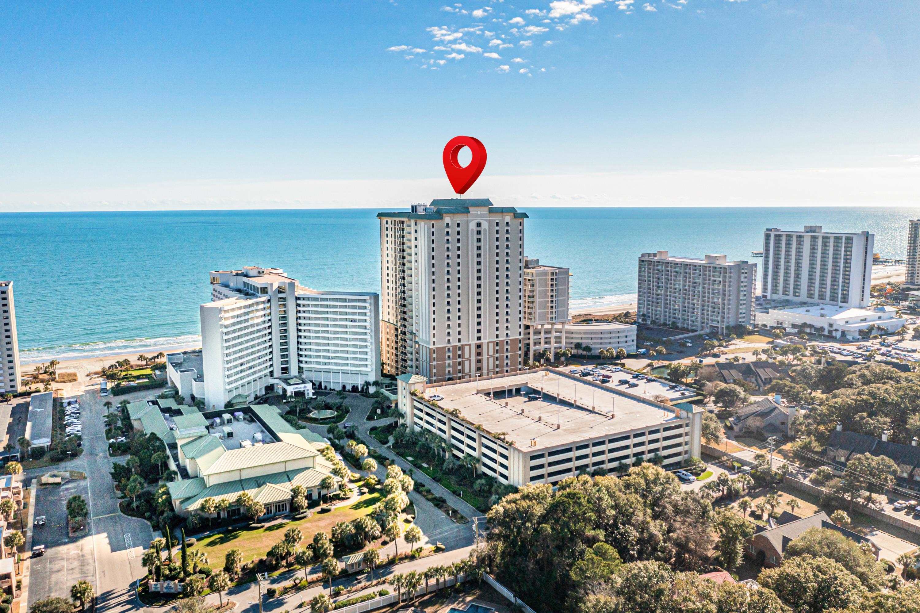 Beachfront condominium tower marked by a red pin, with the ocean and neighboring high-rises behind it.