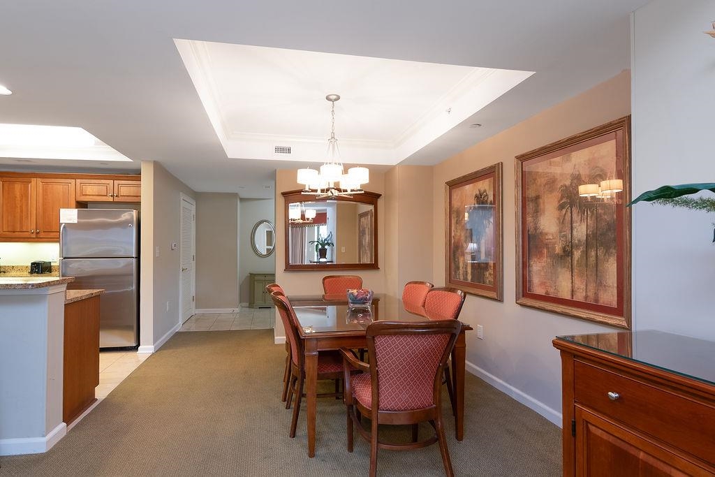 Dining area with a glass-top table, upholstered chairs, chandelier, and adjoining kitchen.