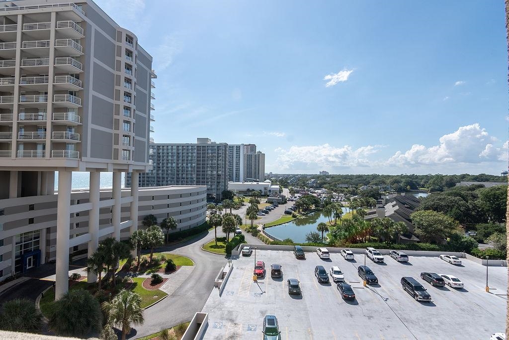 Coastal condominium building with balconies overlooking a parking lot, pond, and distant ocean.