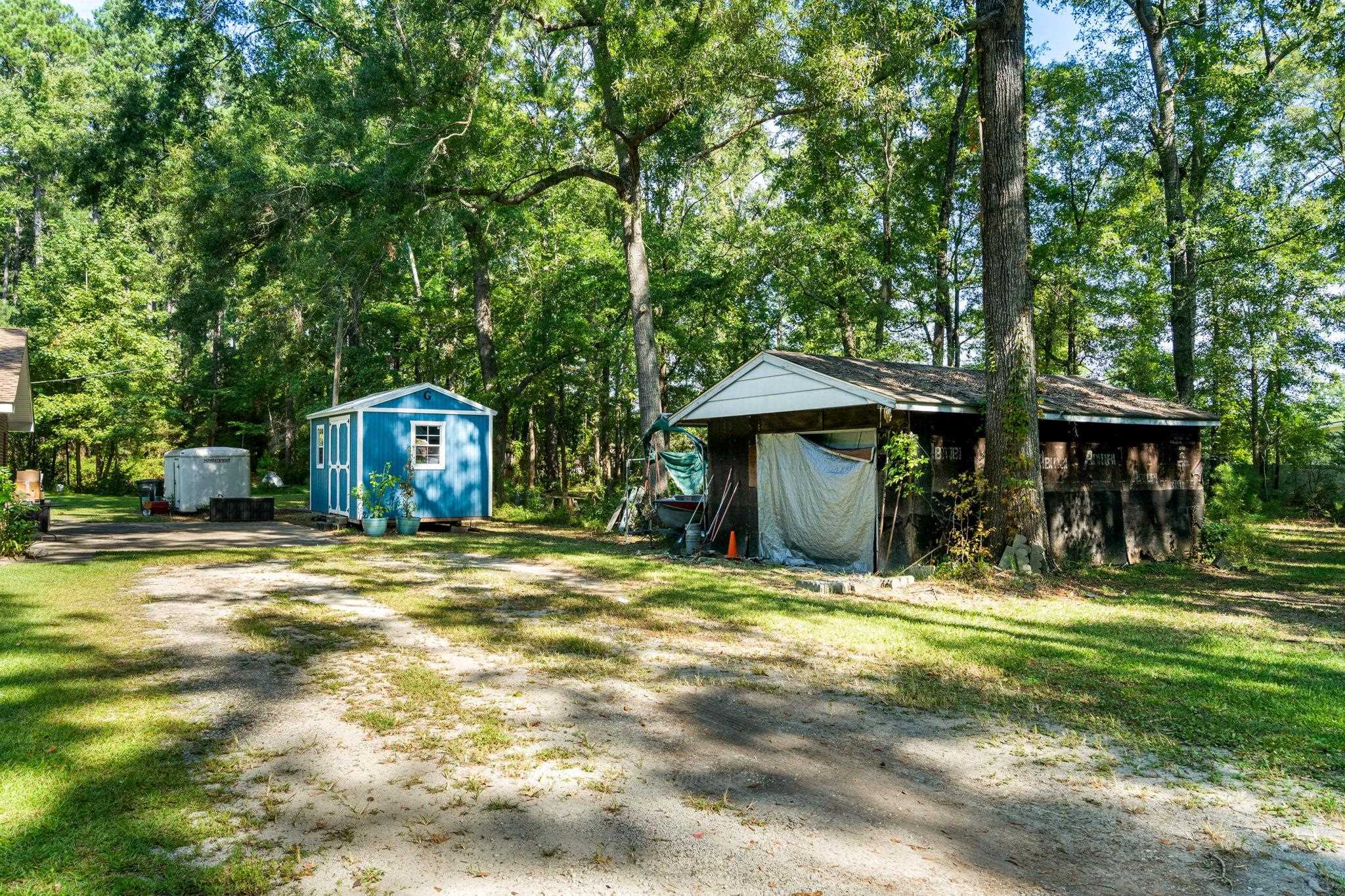 Wooded property yard with a blue shed, covered storage structure, trailer, and sandy driveway.