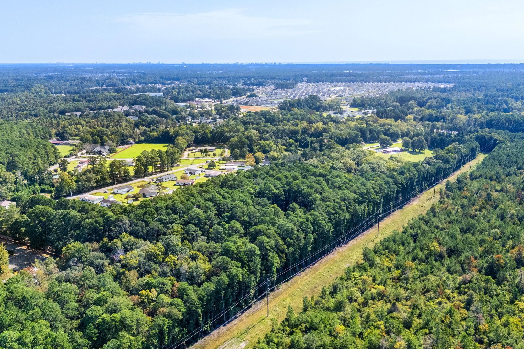 Aerial view of wooded land, scattered homes, and distant neighborhoods beside a power line corridor.