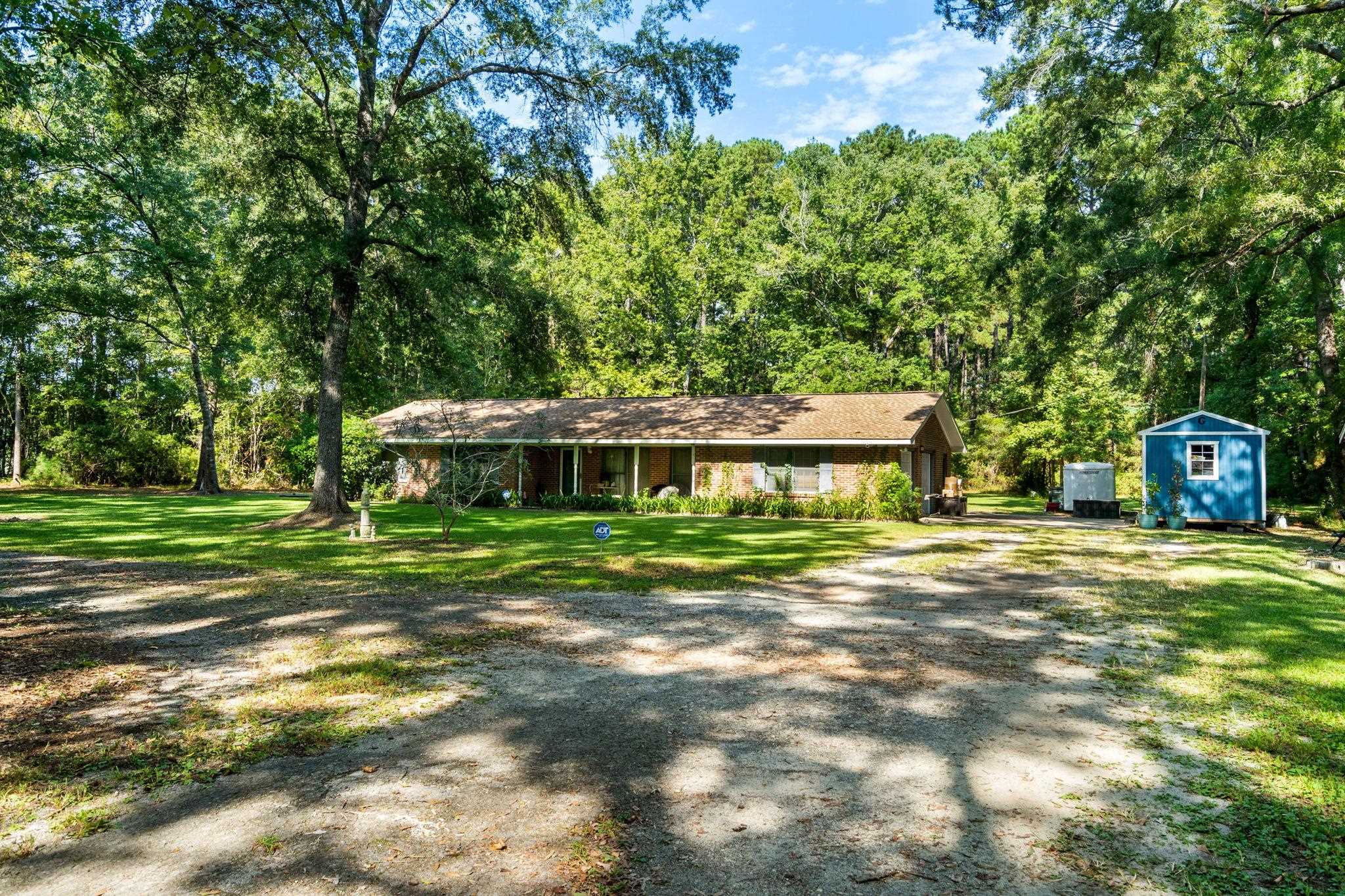 Brick ranch house beneath mature trees, with a gravel driveway and blue shed.