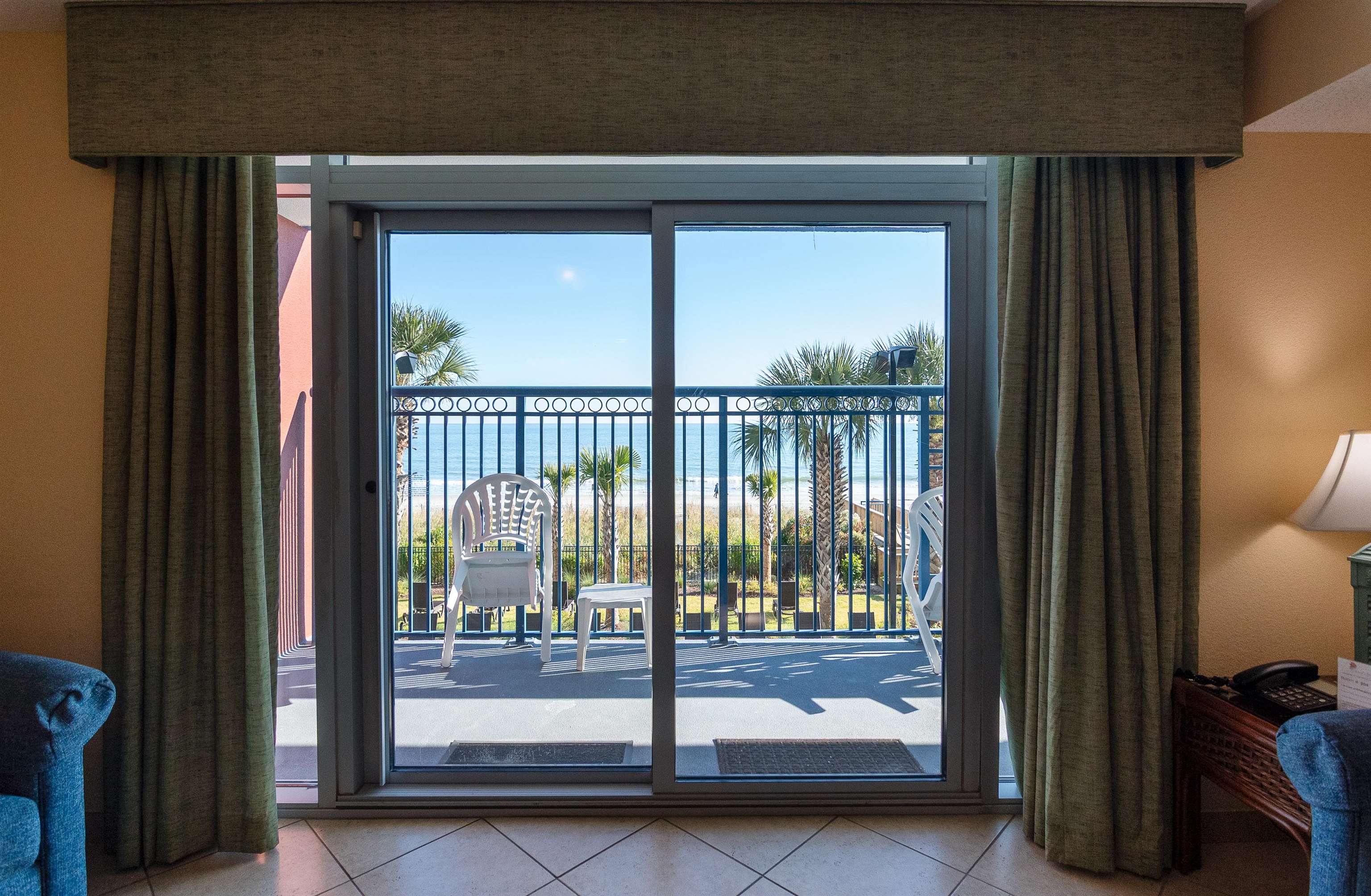 Condo sliding doors open to a balcony with white chairs and a palm-lined ocean view.