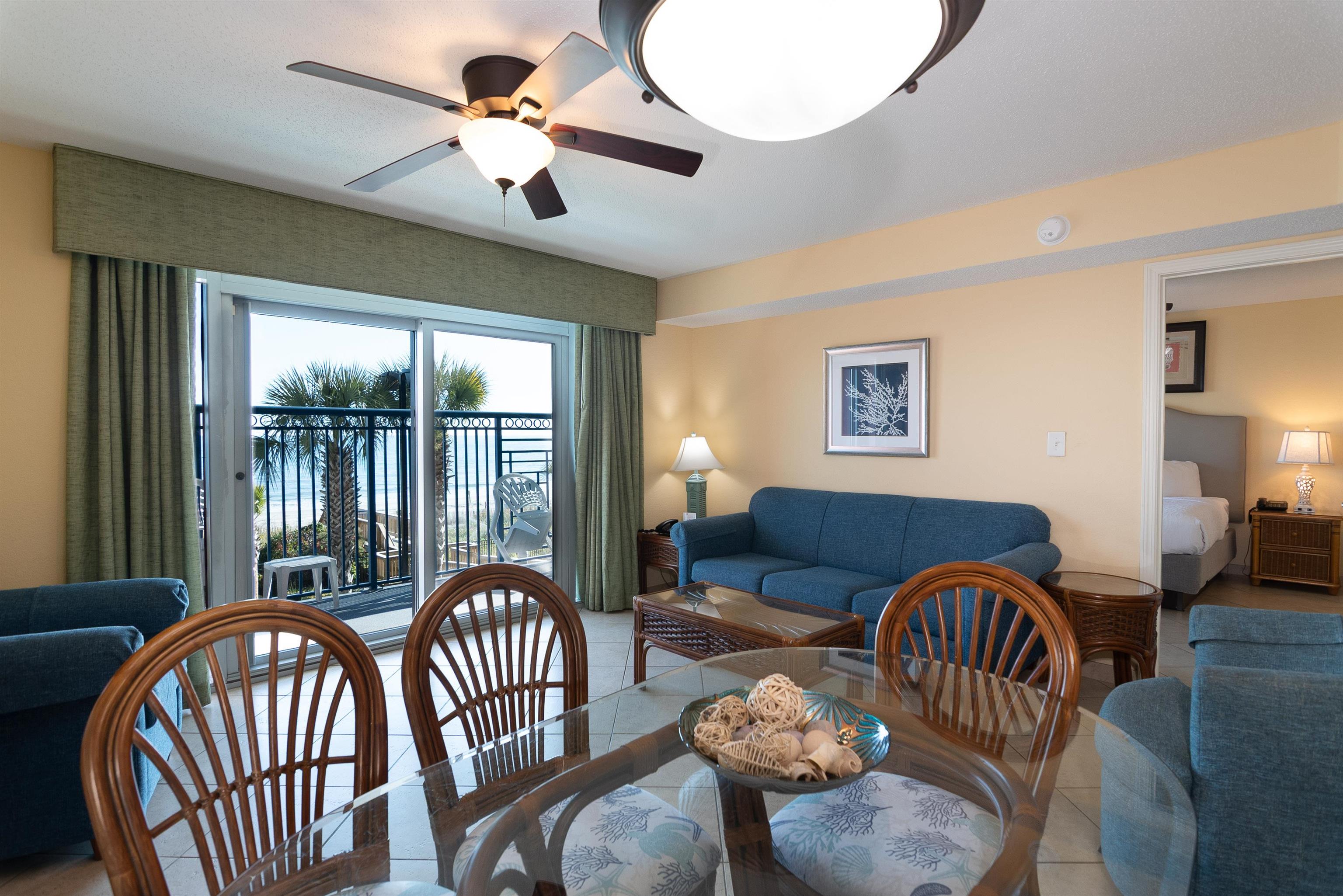 Condo living and dining room with blue sofas and a glass table, opening to a beachfront balcony.