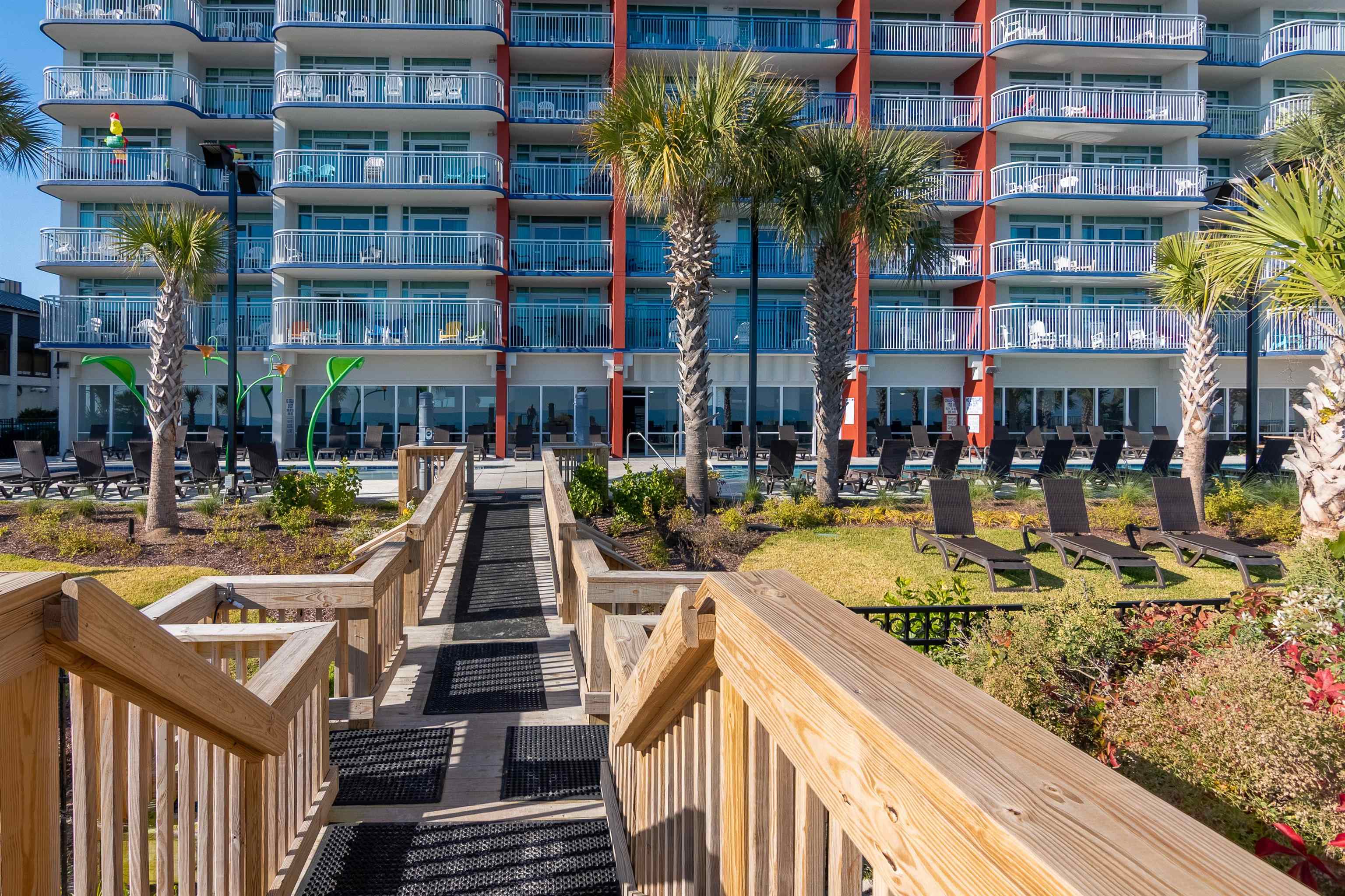 Condominium pool deck with palm trees, lounge chairs, and a wooden ramp to the building.
