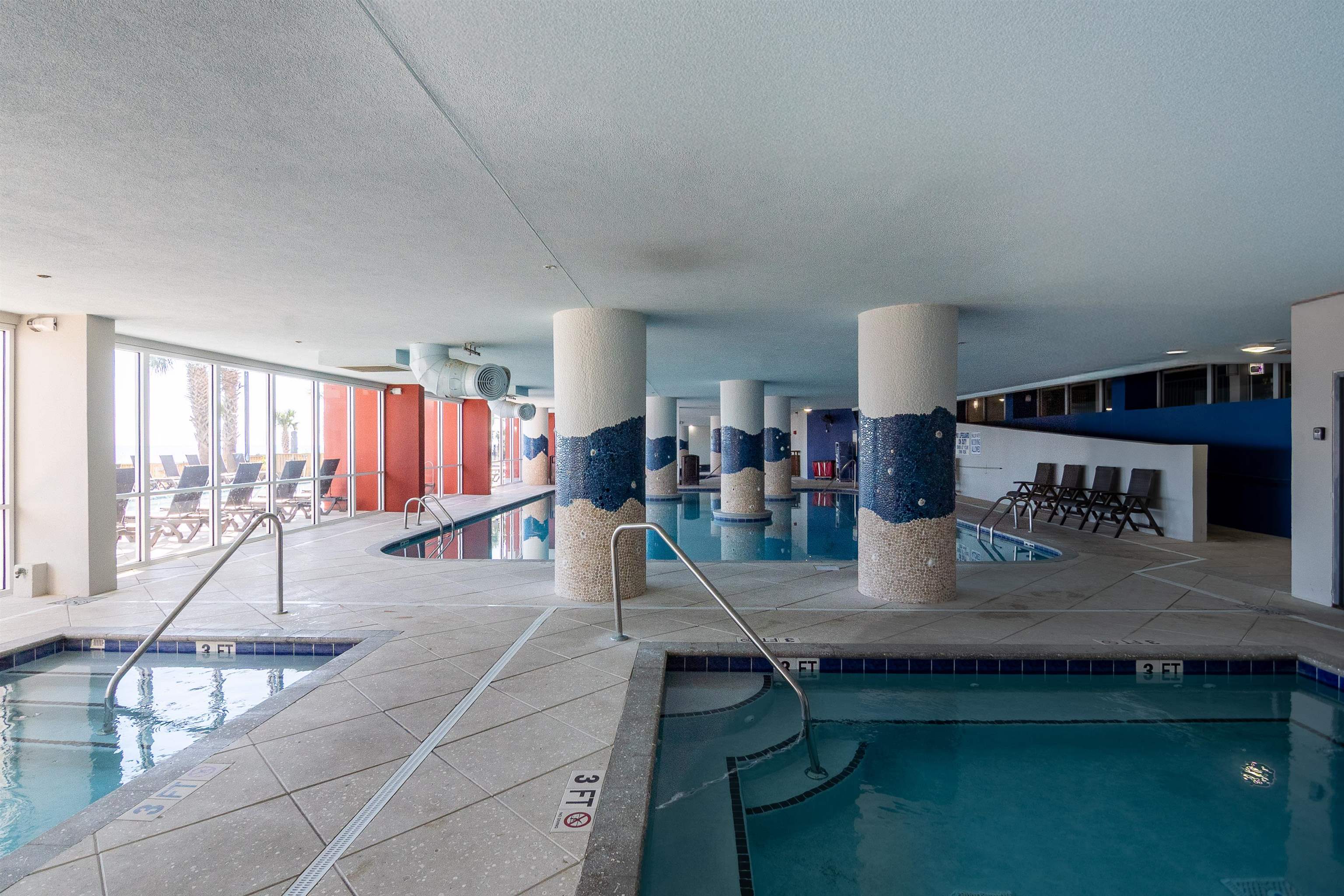 Indoor swimming pool with mosaic-tiled columns and large windows overlooking poolside lounge chairs.
