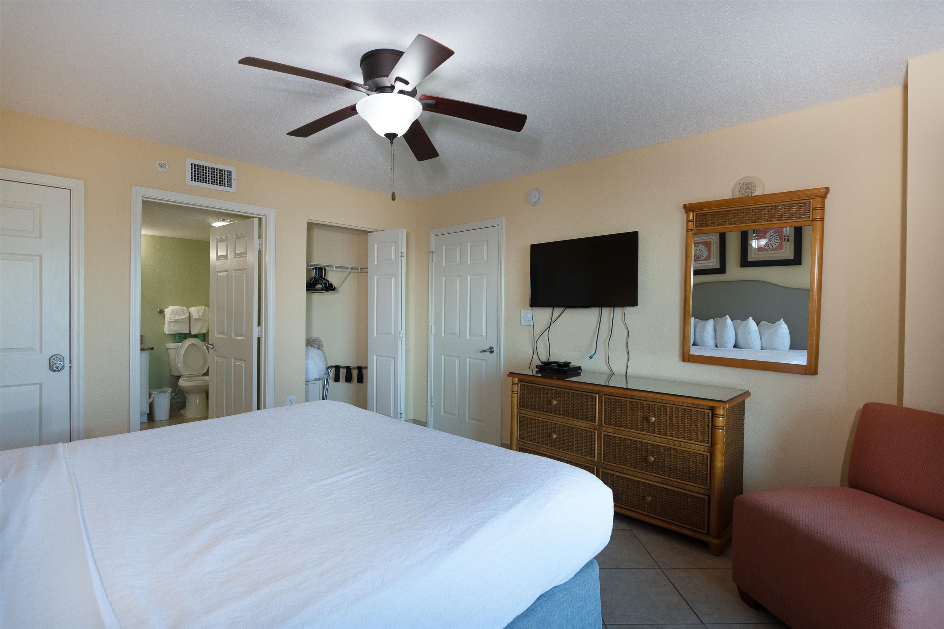 Furnished bedroom with a white bed, ceiling fan, and wicker dresser beneath a wall-mounted television.