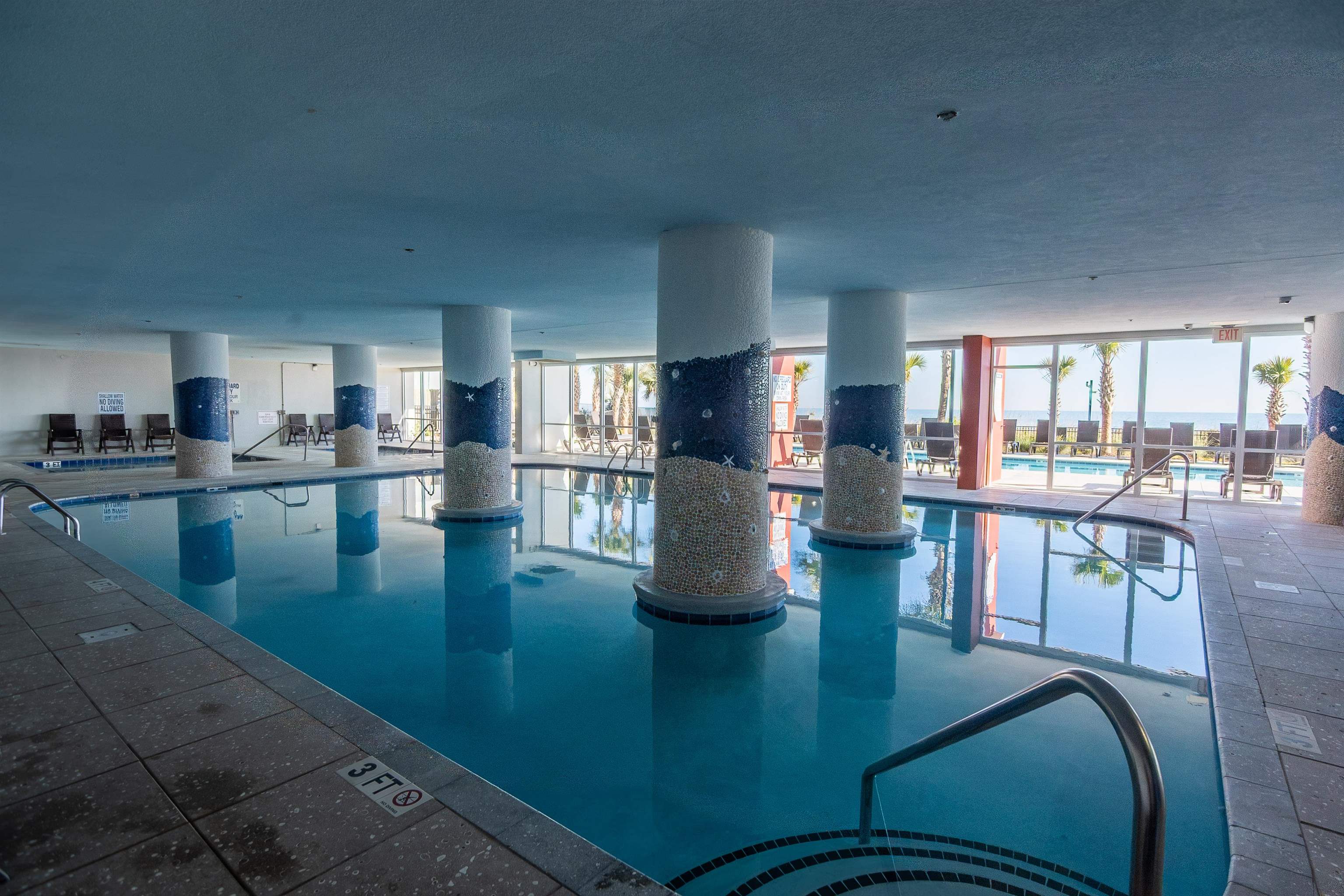 Indoor swimming pool with mosaic columns and windows overlooking palm trees and the ocean.
