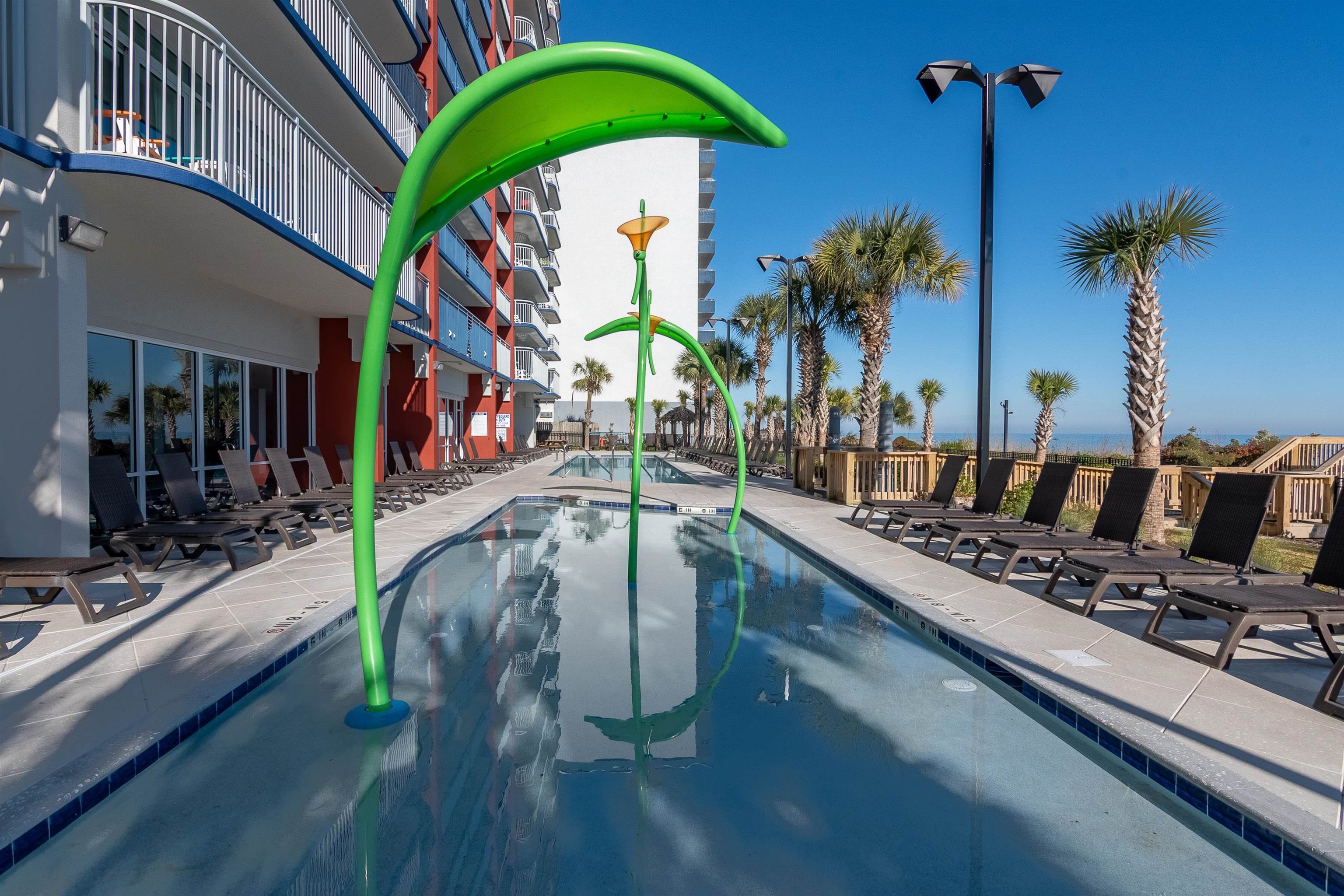 Condominium pool with a large green flower-shaped water feature, lounge chairs, palm trees, and ocean views.