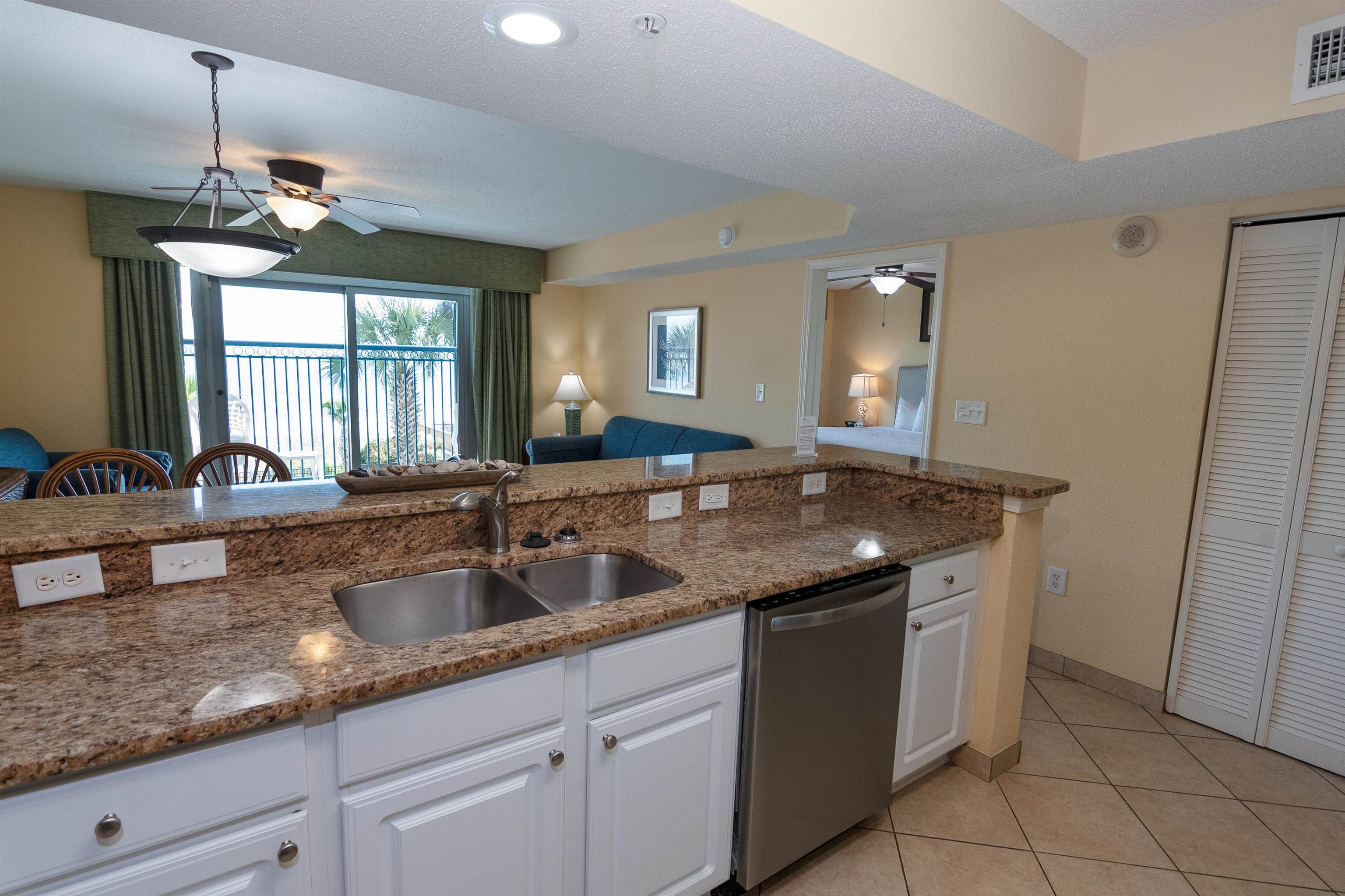 Condo kitchen with granite counters and stainless dishwasher, opening to a balcony with water and palm trees.