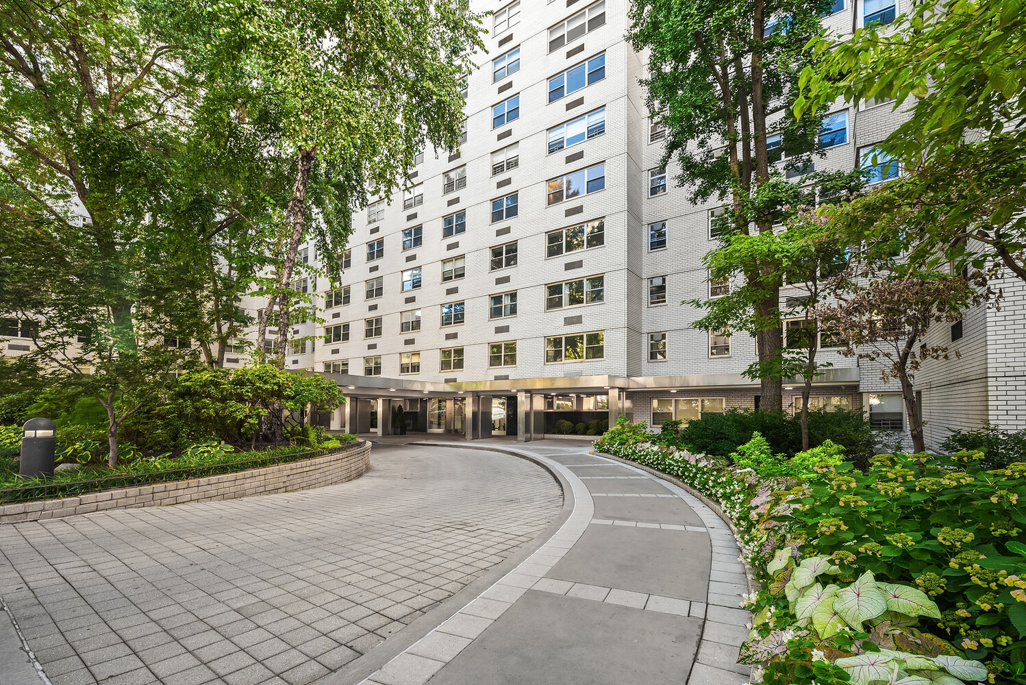 White brick apartment building behind a curved driveway bordered by trees and flower beds.