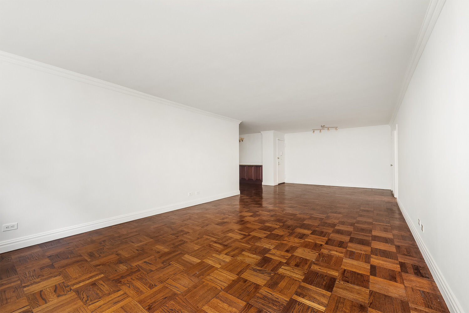 Vacant living room with patterned parquet floors, white walls, and a glimpse of dark kitchen cabinetry.