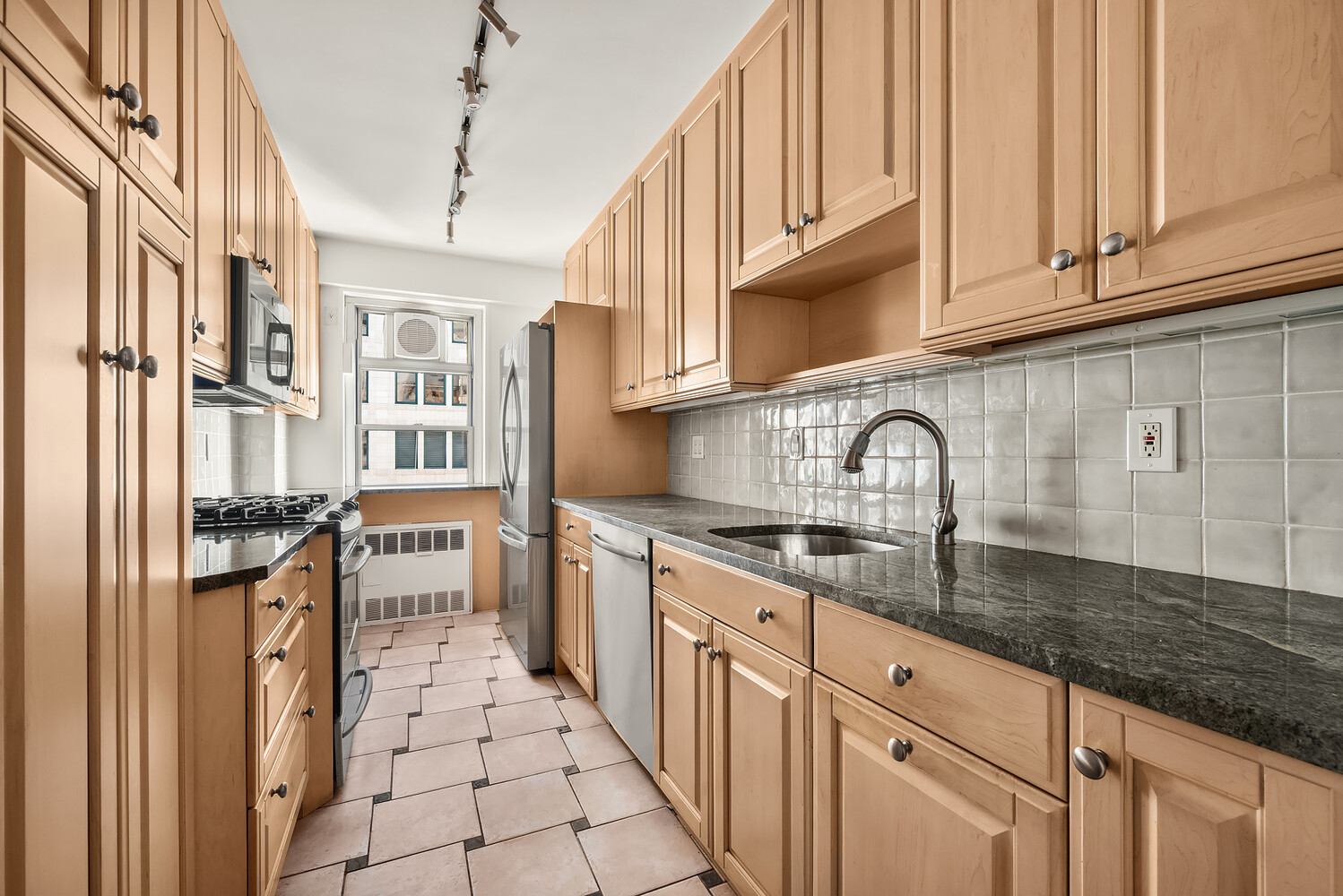 Galley kitchen with light wood cabinets, dark countertops, and a window at the far end.