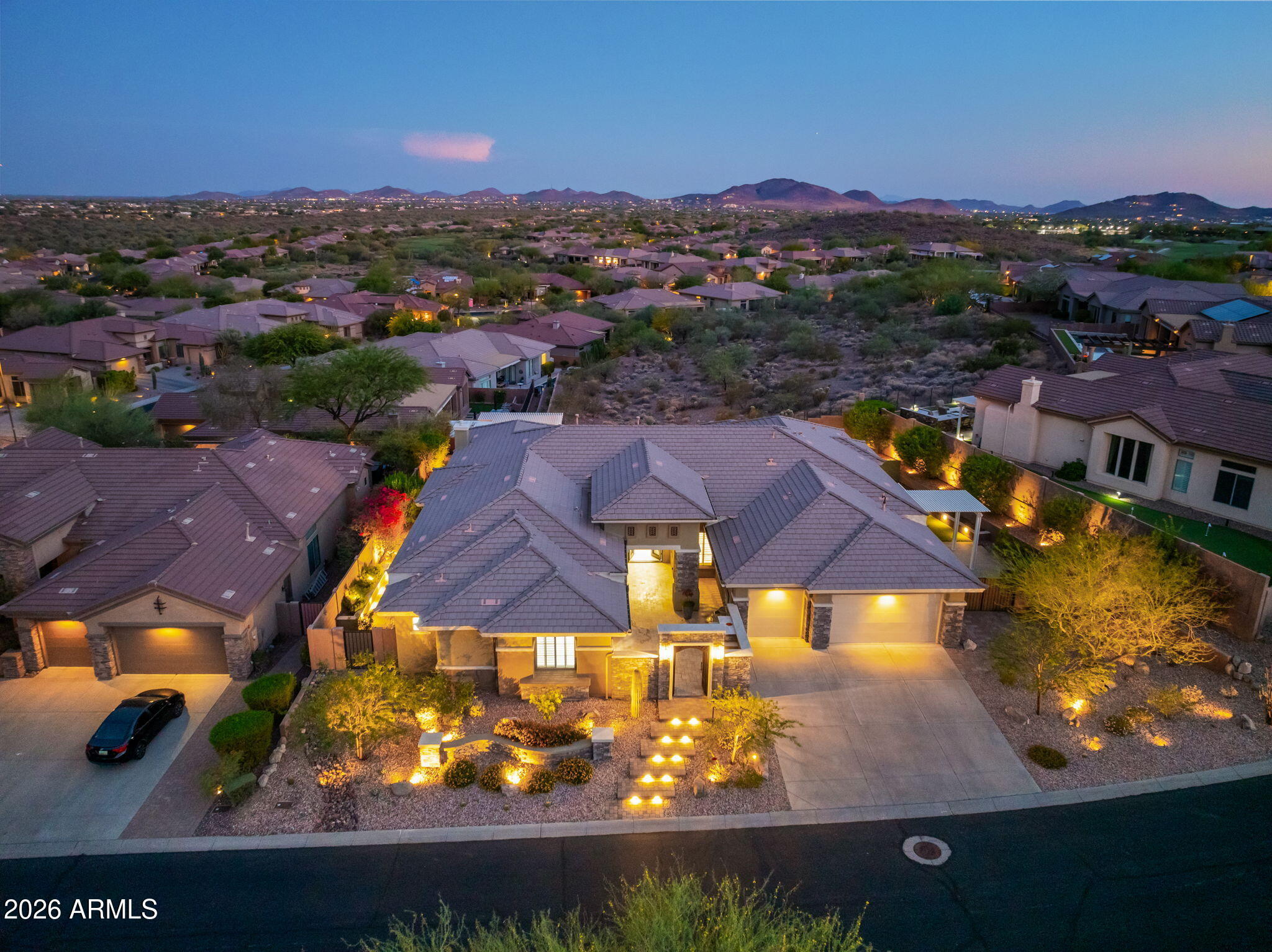 Aerial view of a tile-roof home with a lit entry and three-car garage, surrounded by desert homes and mountains at dusk.