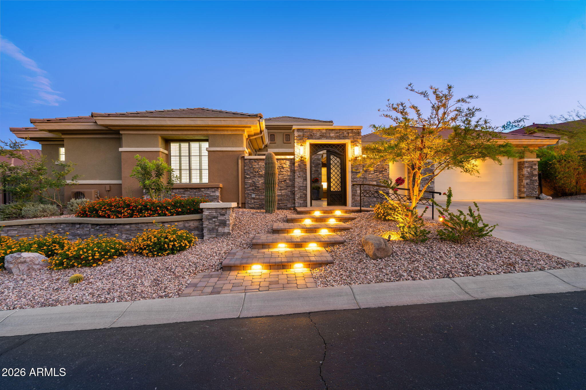 Single-story stucco home with stone accents, illuminated entry steps, and desert landscaping at dusk.