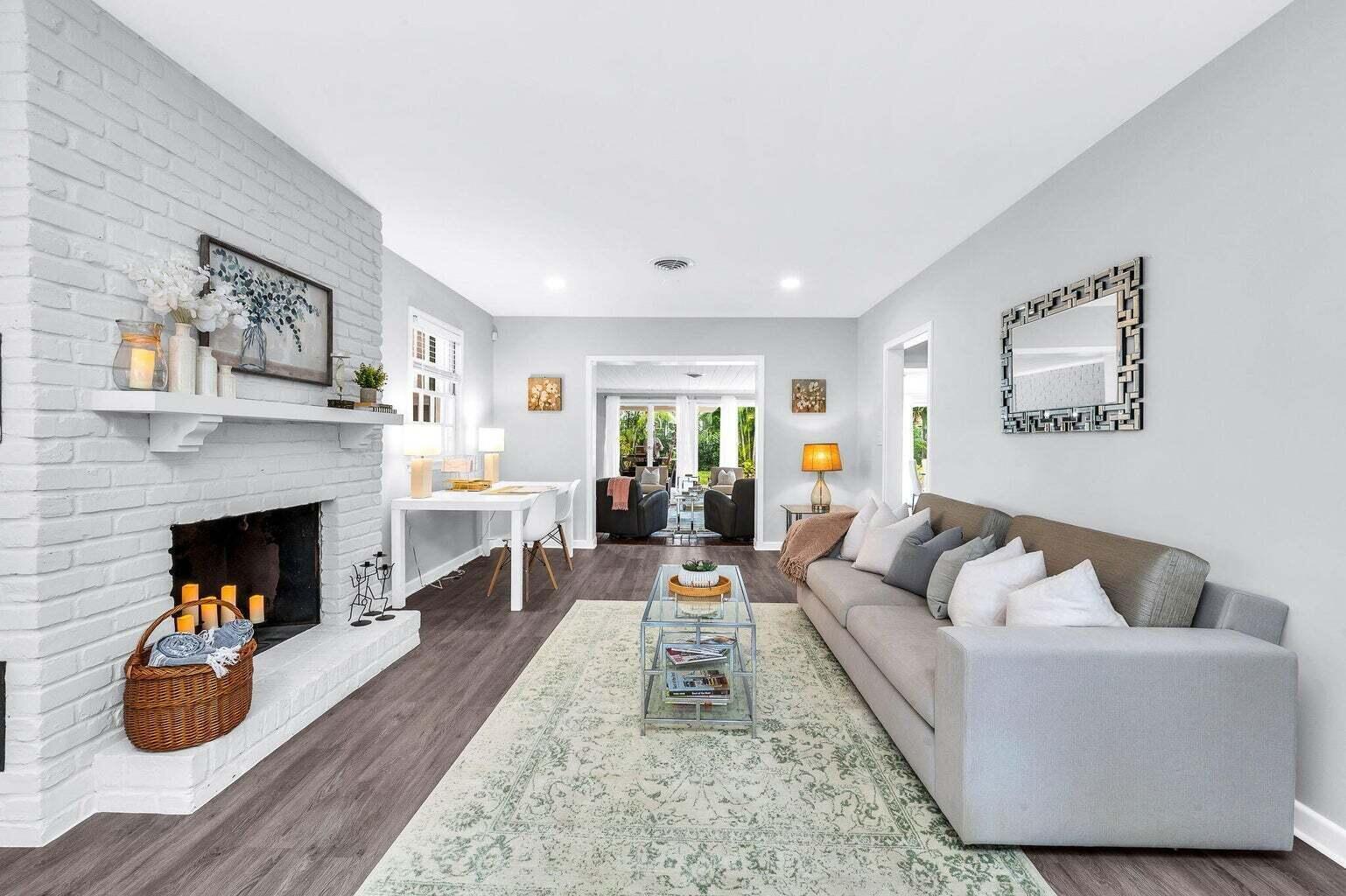 Staged living room with a white brick fireplace, gray sectional, and doorway to a bright sitting area.