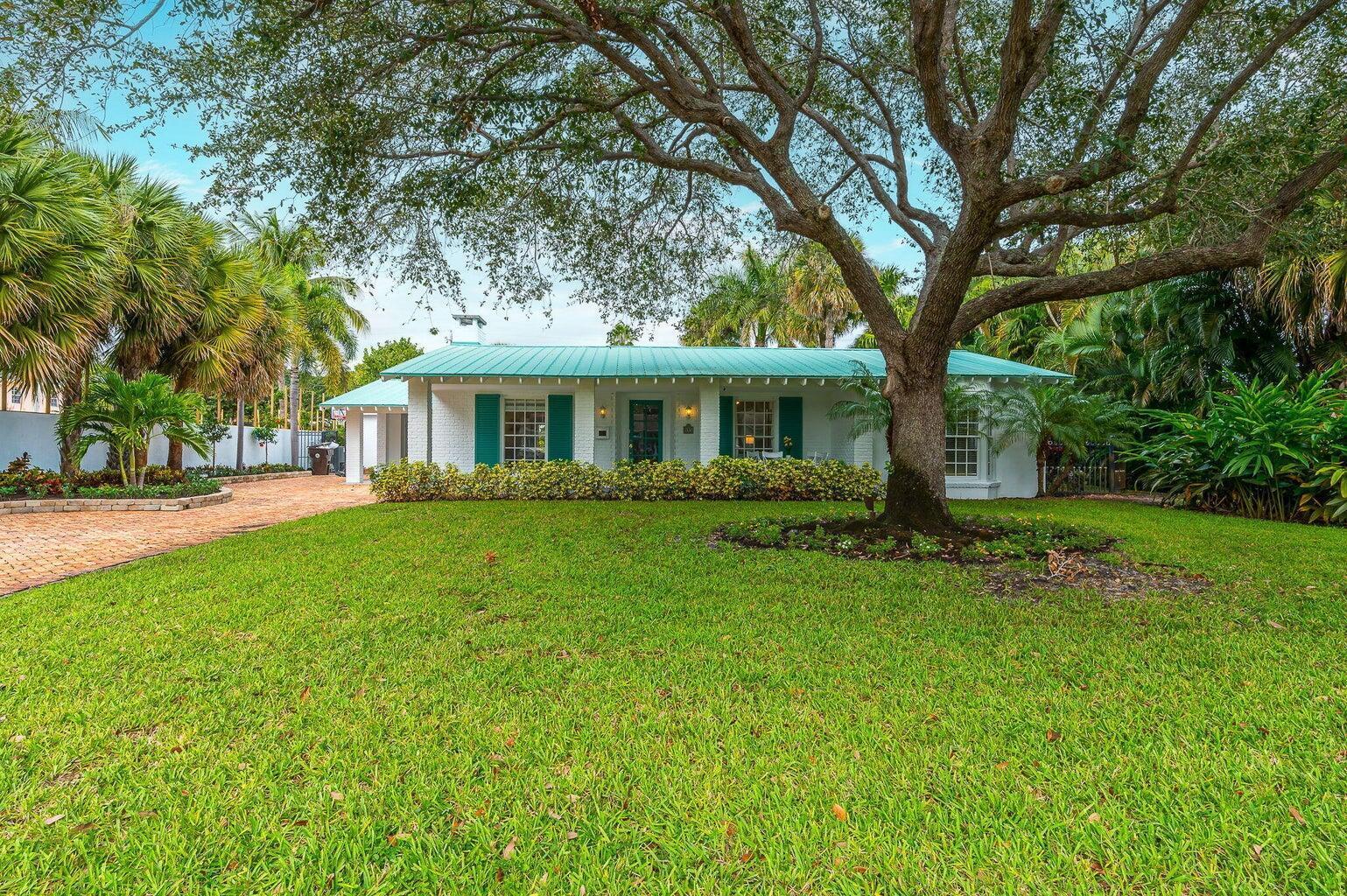 White single-story house with teal roof and shutters, shaded by a large oak beside a brick driveway.