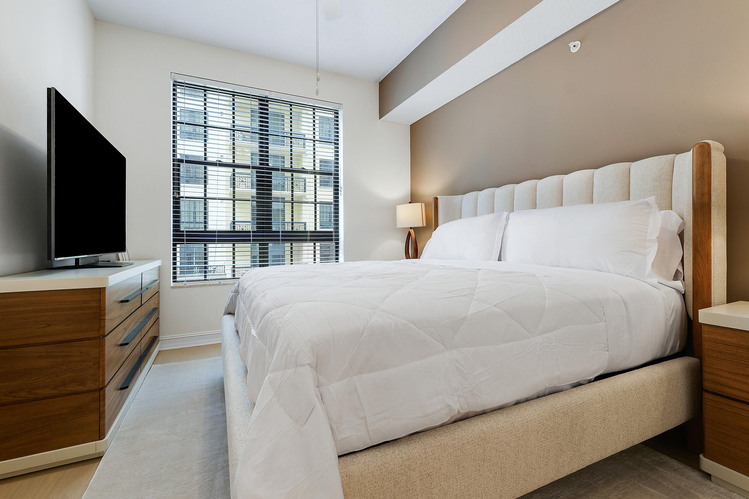 Bedroom with a large white bed, wood furniture, and a window overlooking a neighboring building.