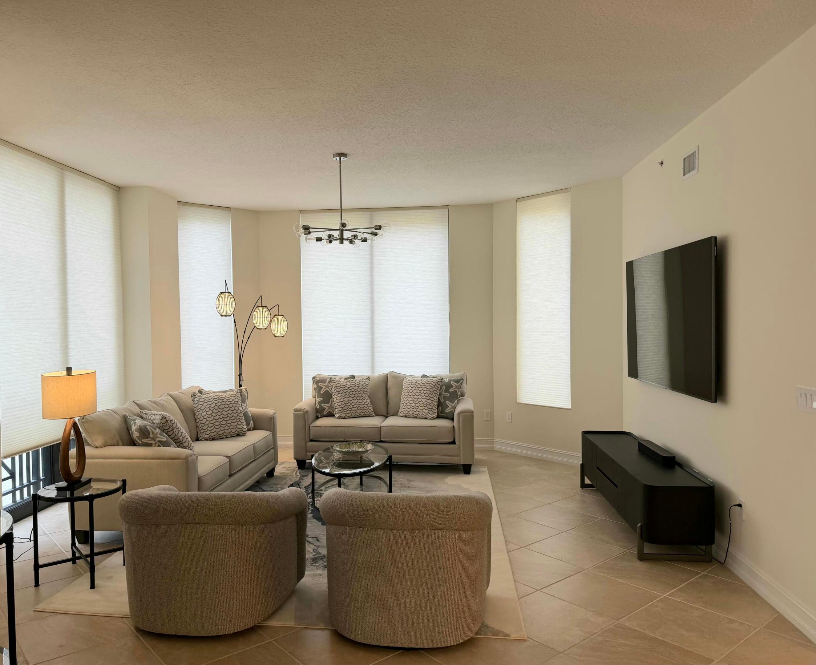 Furnished living room with beige seating, tall shaded windows, and a wall-mounted television.