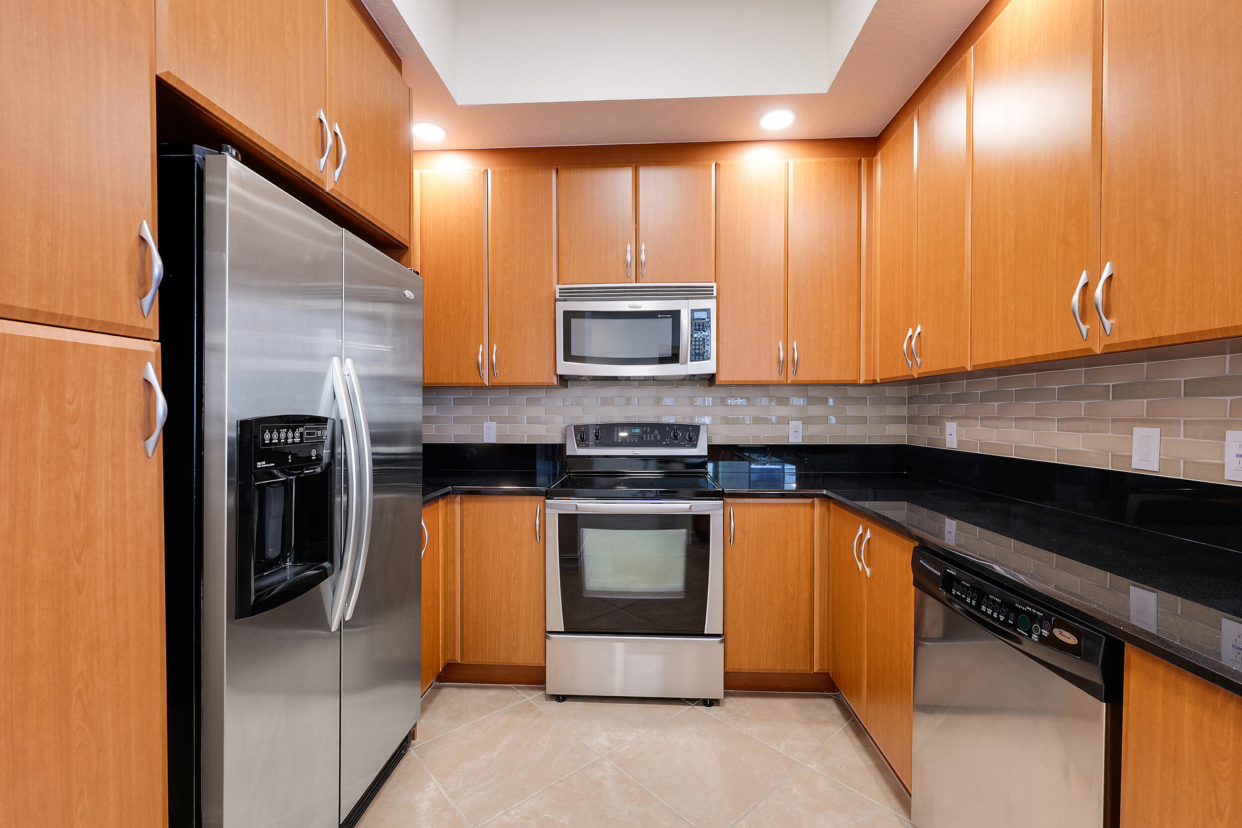 Kitchen with warm wood cabinets, black countertops, gray tile backsplash, and stainless-steel appliances.
