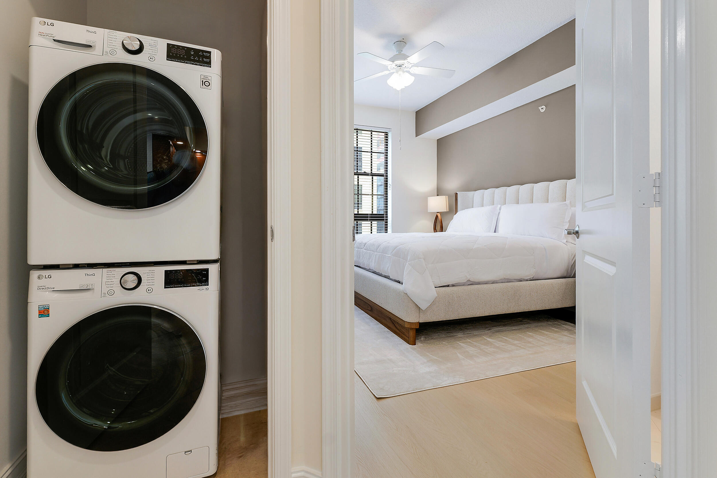 Staged bedroom viewed past a stacked washer and dryer, with a white bed and ceiling fan.