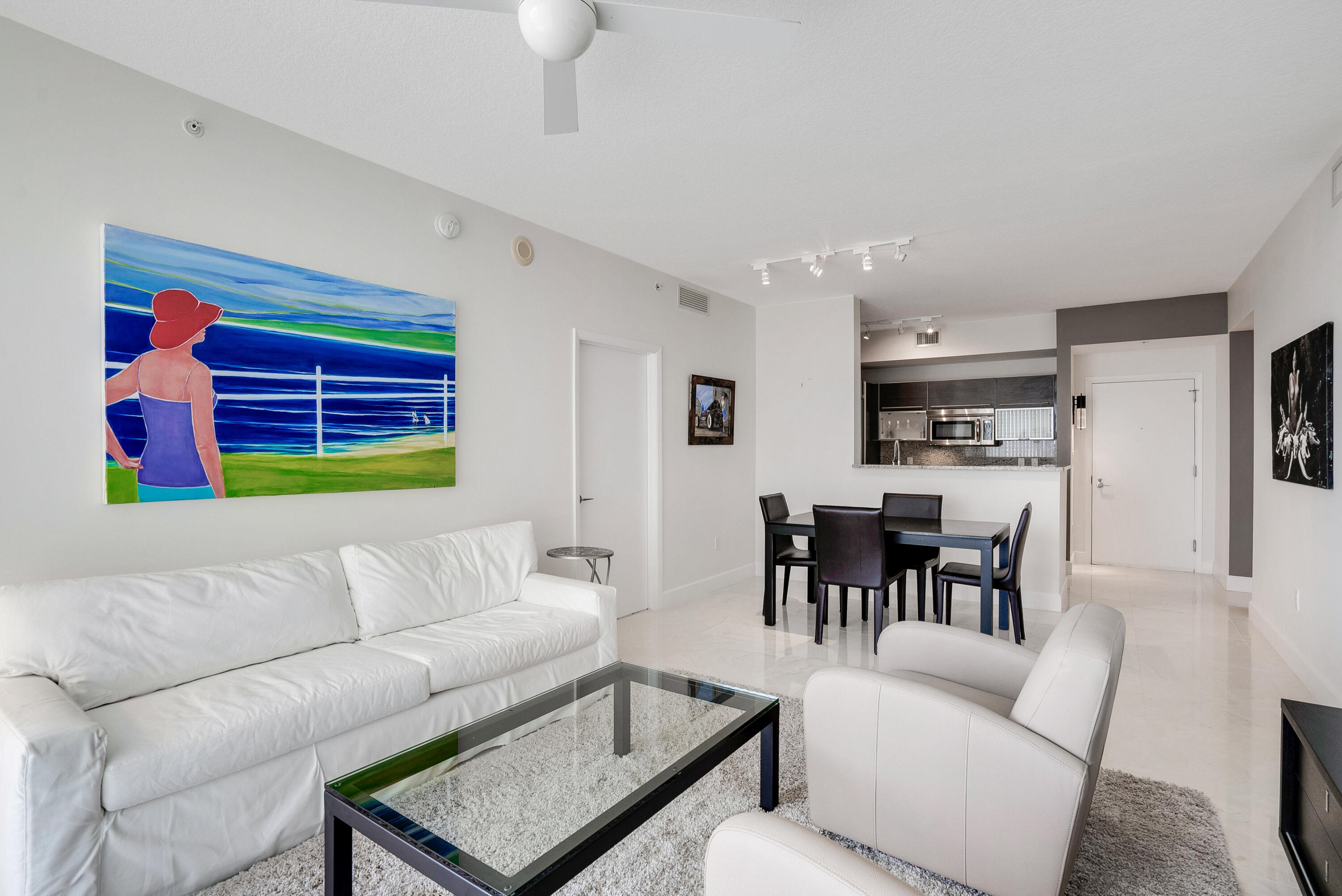 Furnished condo living room with white seating, colorful beach artwork, and an open dining area and kitchen.