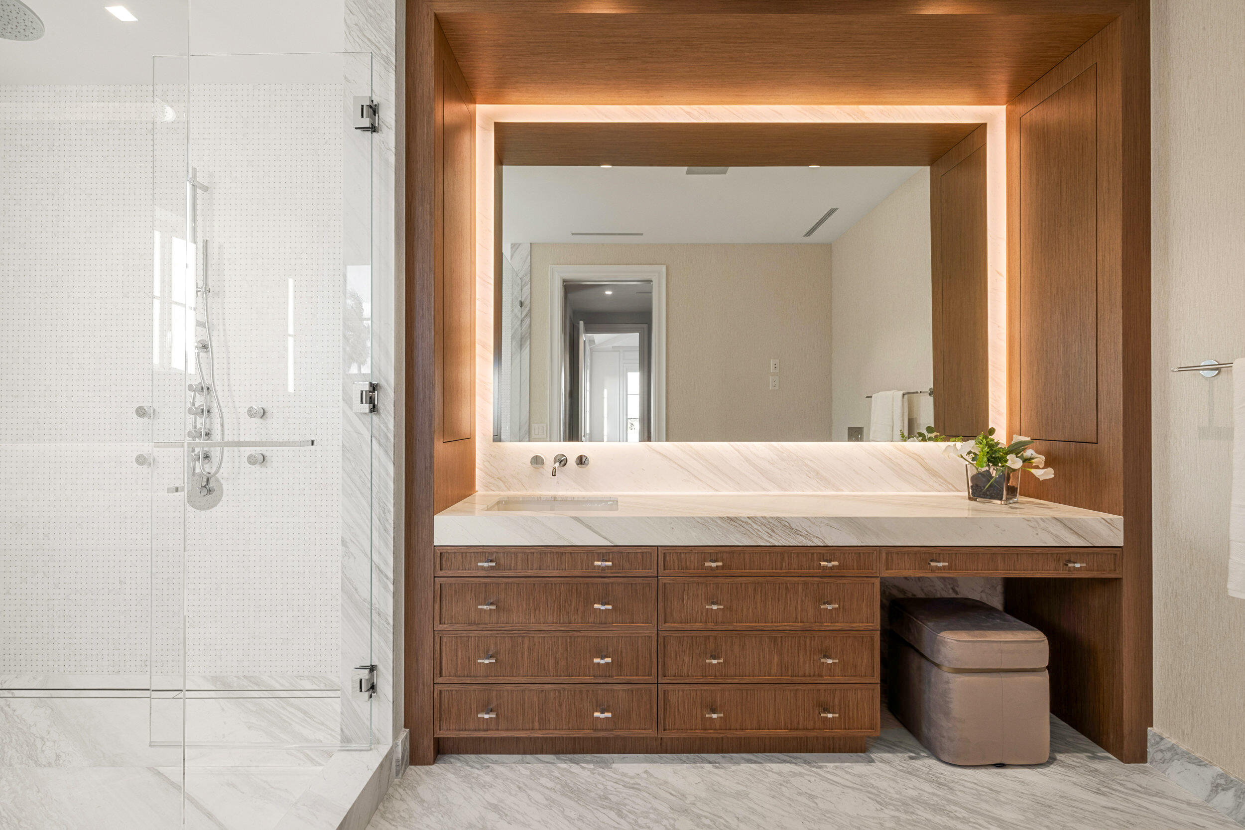 Modern bathroom with a glass marble shower and walnut vanity framed by a warm backlit mirror.