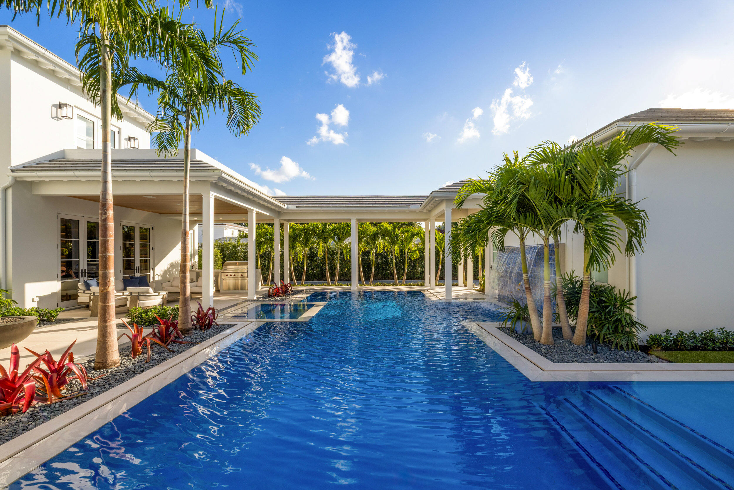 Swimming pool beside a covered patio with lounge seating, a built-in grill, and tropical palms.