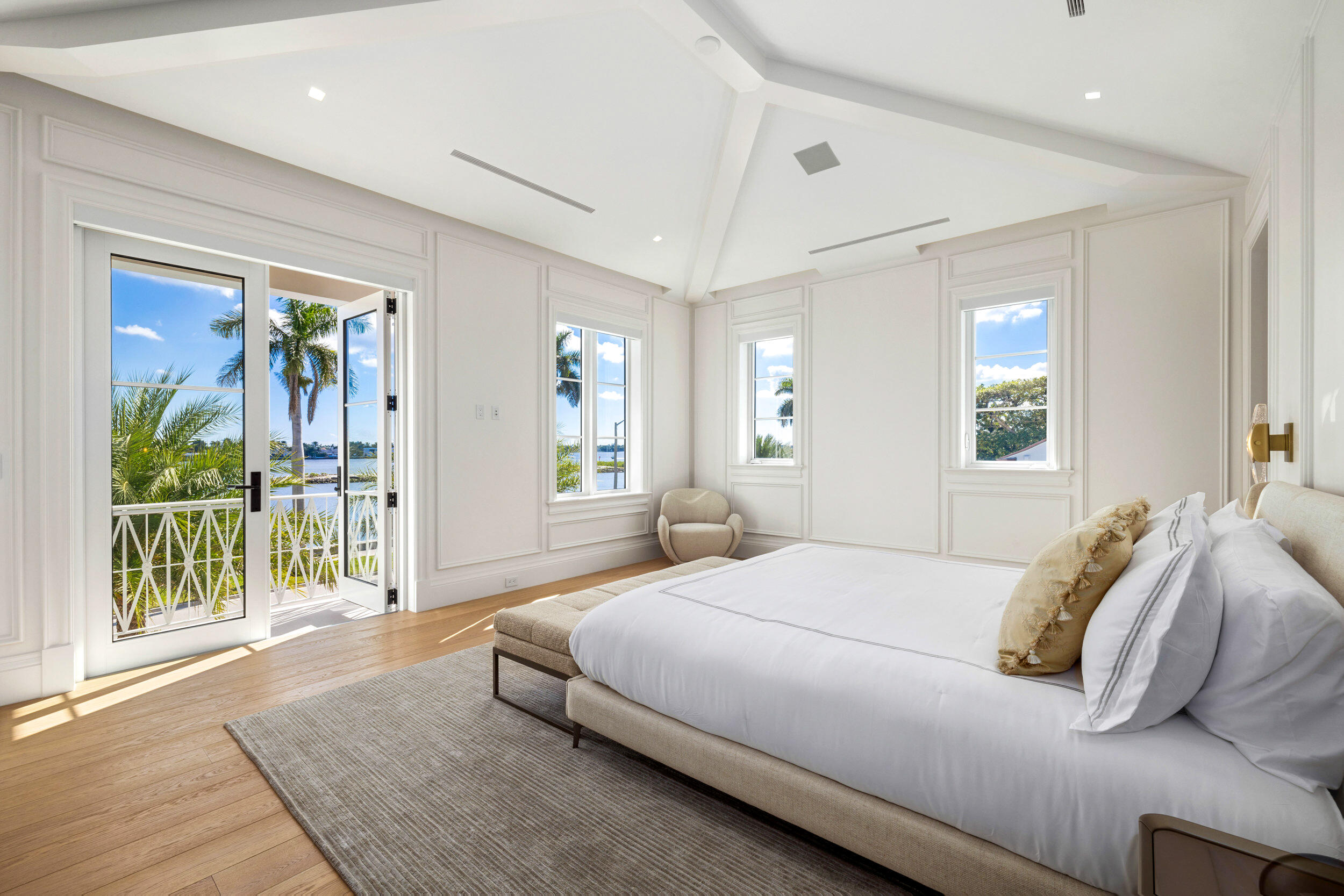 Bright bedroom with a large white bed and balcony doors overlooking palm trees and water.