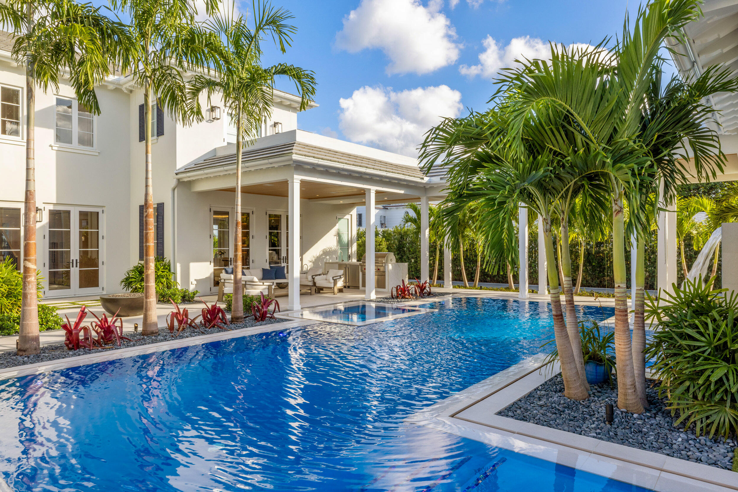 White two-story home behind a blue pool, framed by palm trees and a covered patio.