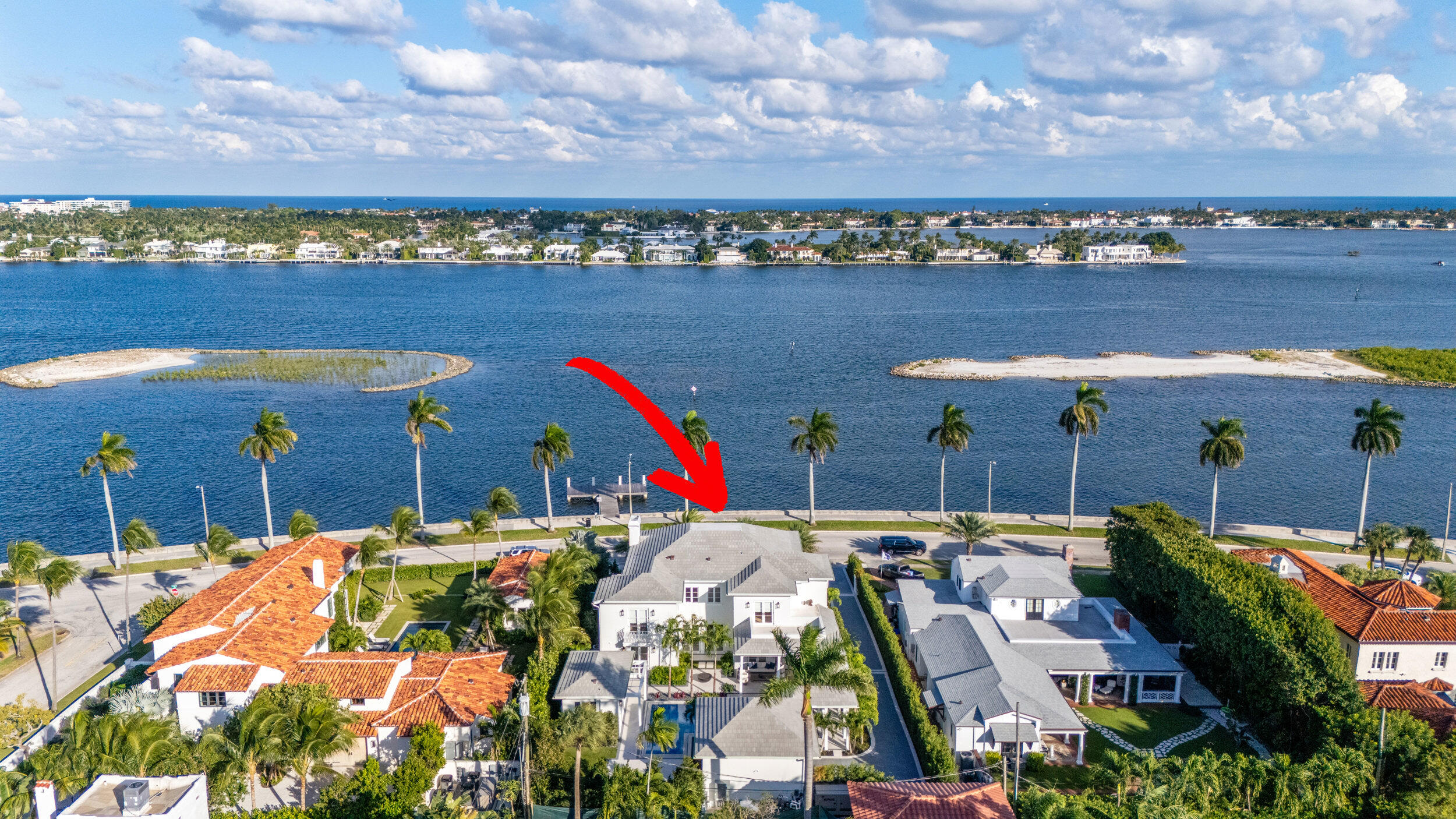 Aerial view of a waterfront home, marked by a red arrow, beside a palm-lined seawall.