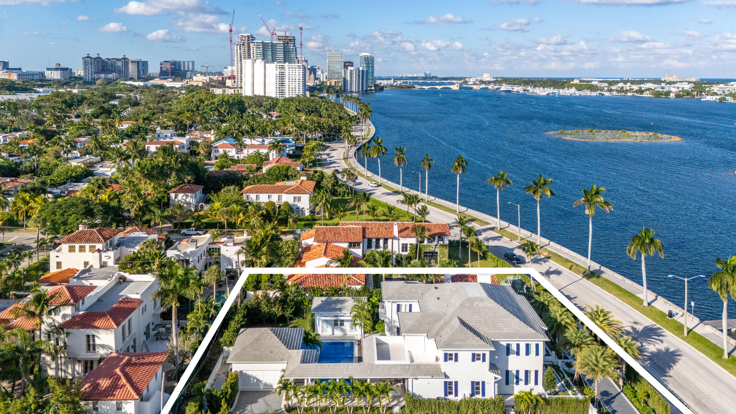 Aerial view of a waterfront home with a pool, white boundary outline, and palm-lined road beside the water.