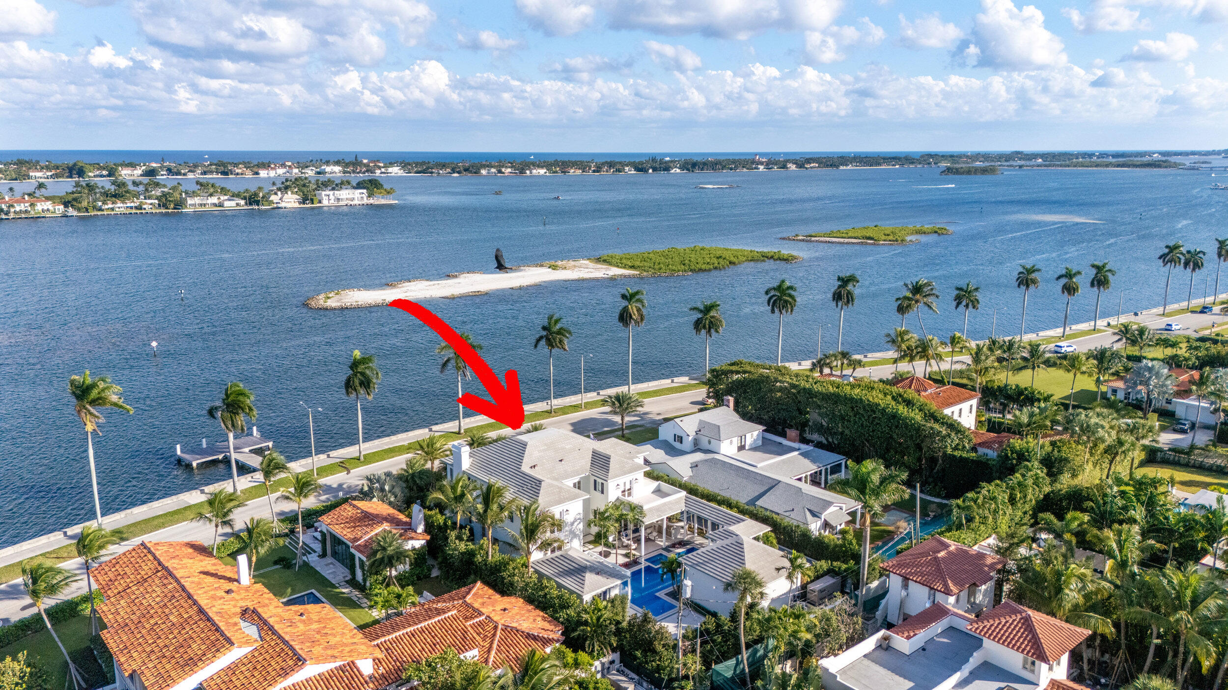 Aerial view of the waterfront home, marked by a red arrow, with a pool and palm-lined shoreline.