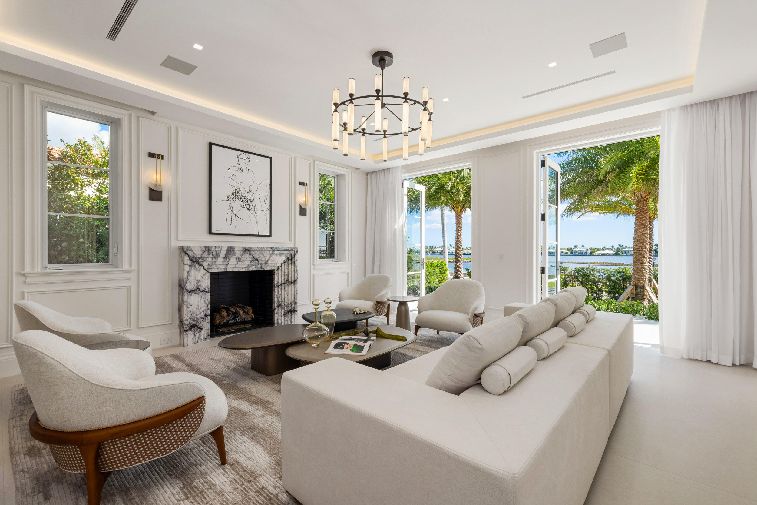 Living room with a marble fireplace and open doors overlooking palm trees and water.