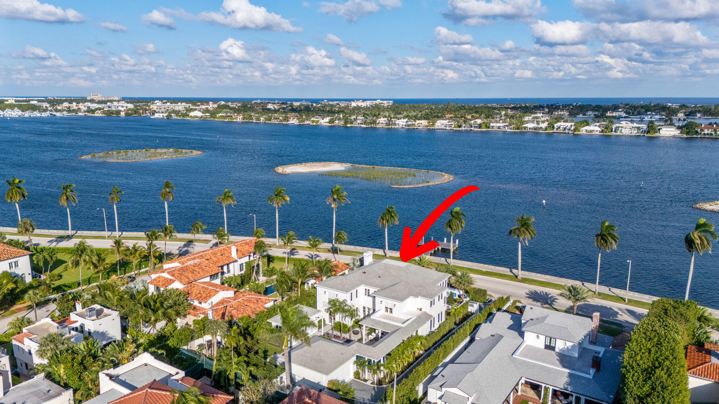 Aerial view of a white waterfront home marked by a red arrow, beside a palm-lined road.