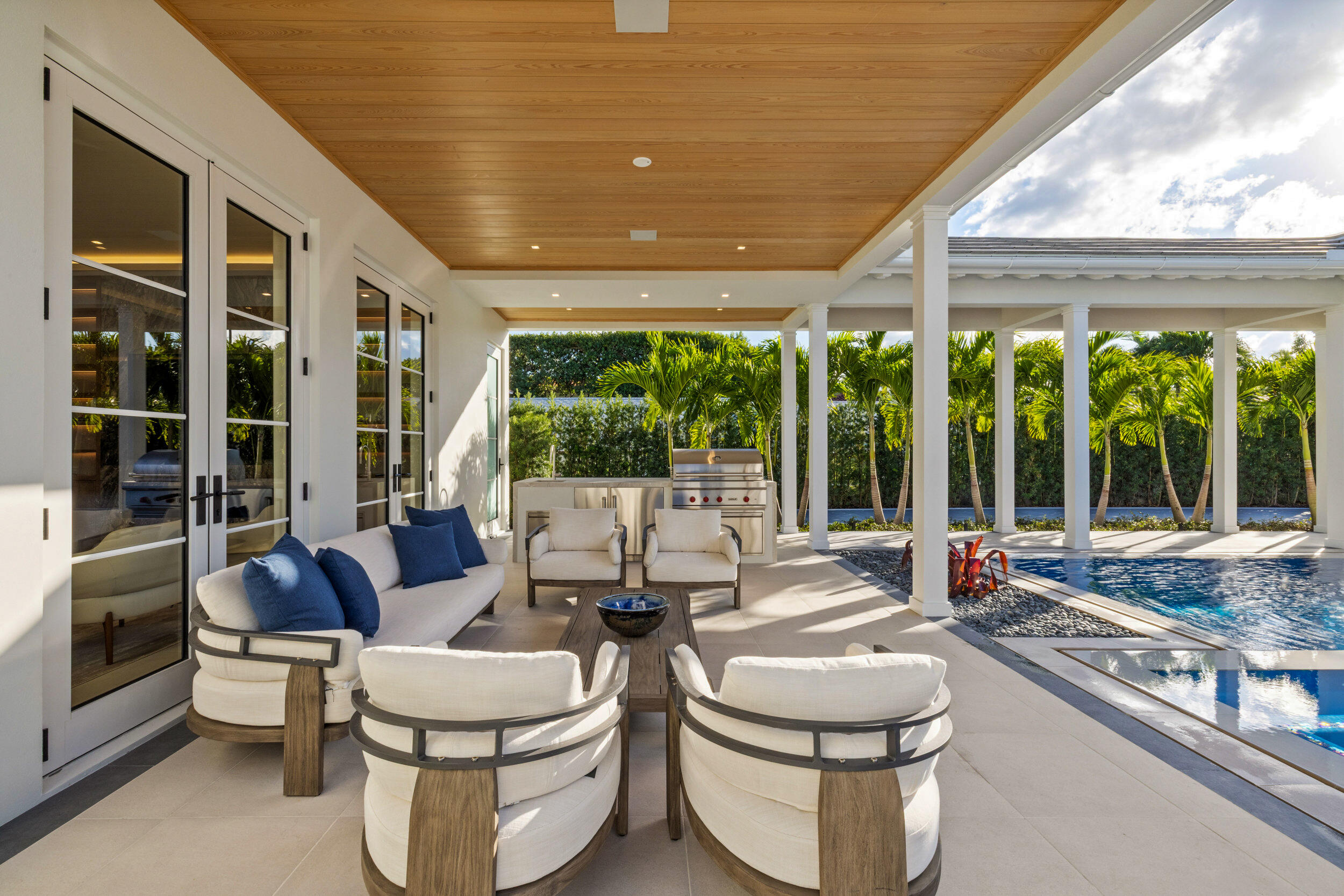 Covered patio with white lounge seating, a built-in grill, and a palm-lined swimming pool.