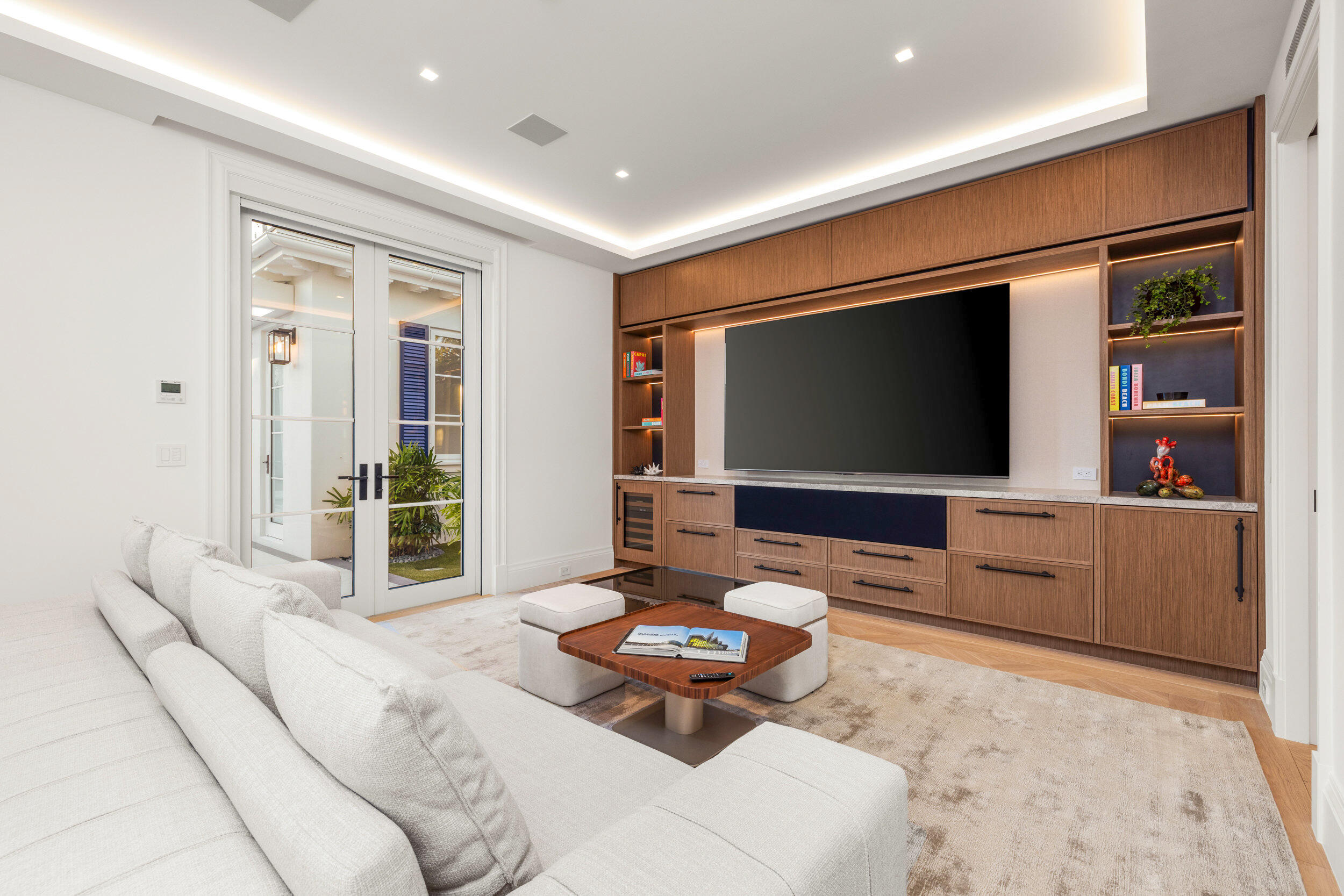 Living room with wood built-ins, a large television, white sectional, and French doors to a planted courtyard.