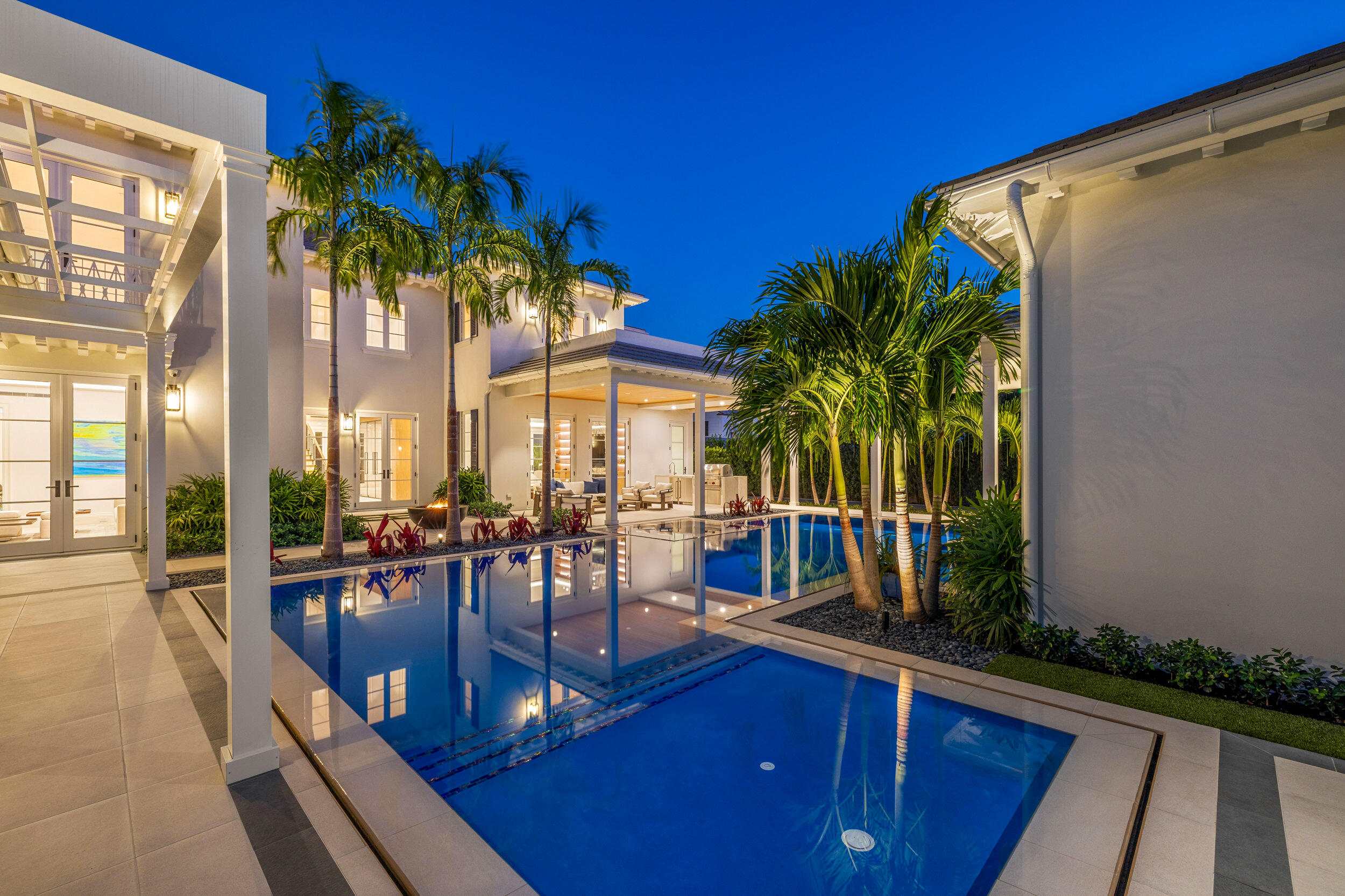 Modern home and palm-lined swimming pool illuminated at dusk, with the house reflected in the water.
