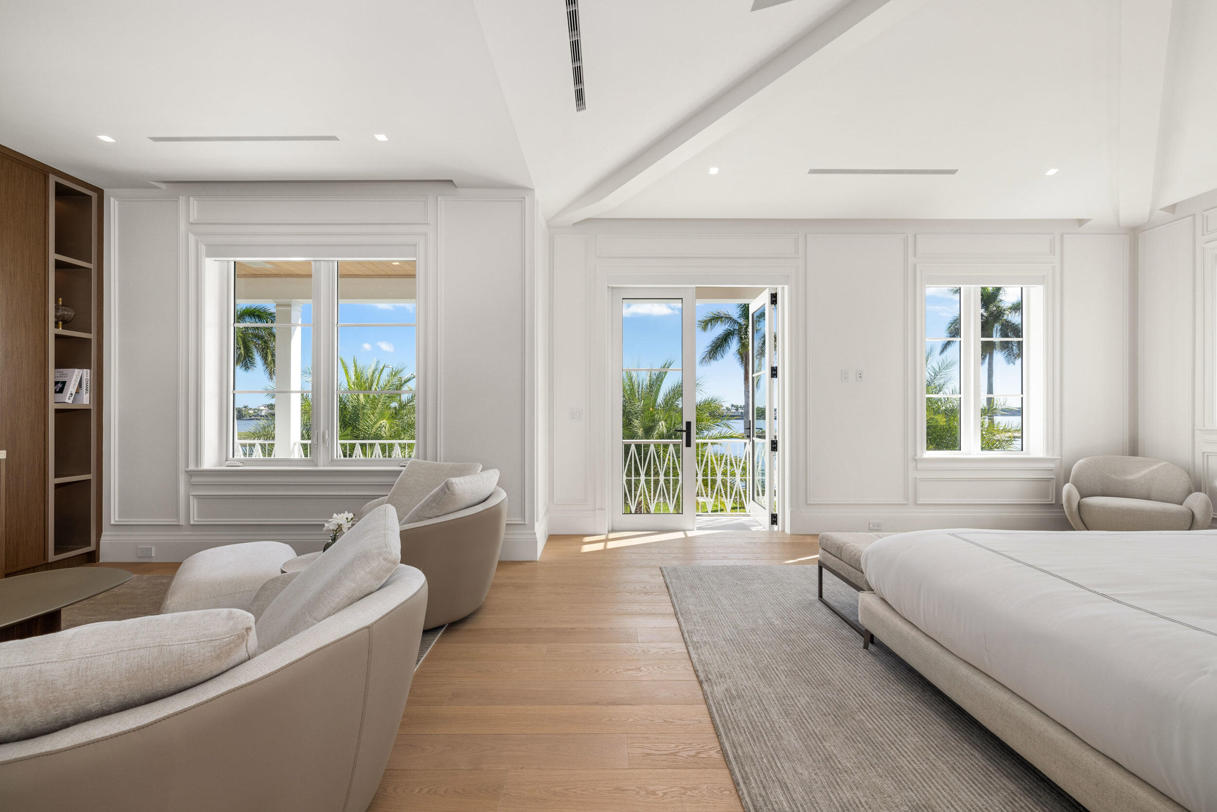 Bright bedroom with a king-size bed, upholstered seating, and open doors overlooking palms and water.