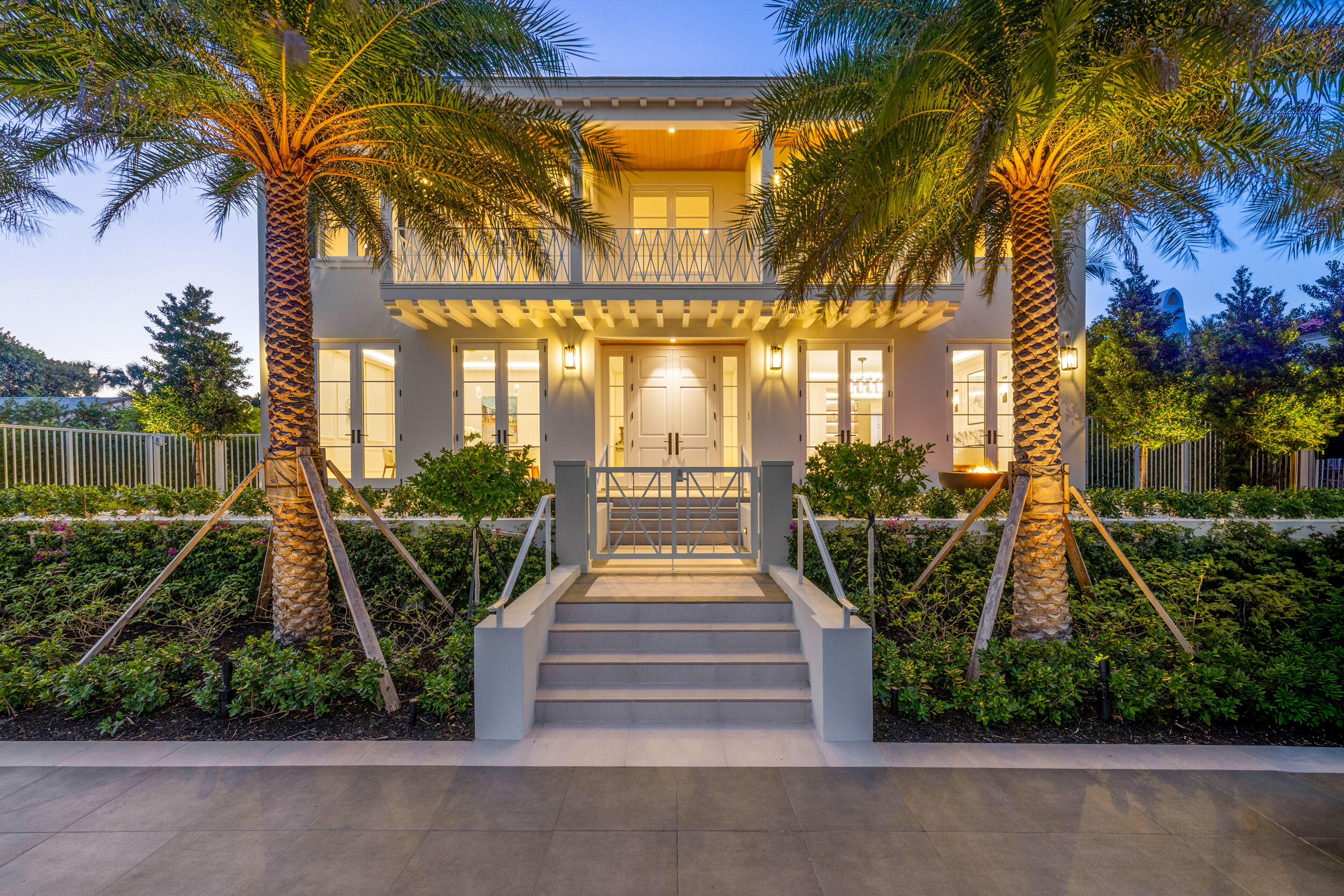 Two-story home entrance with broad steps, balcony, and palm trees at dusk.