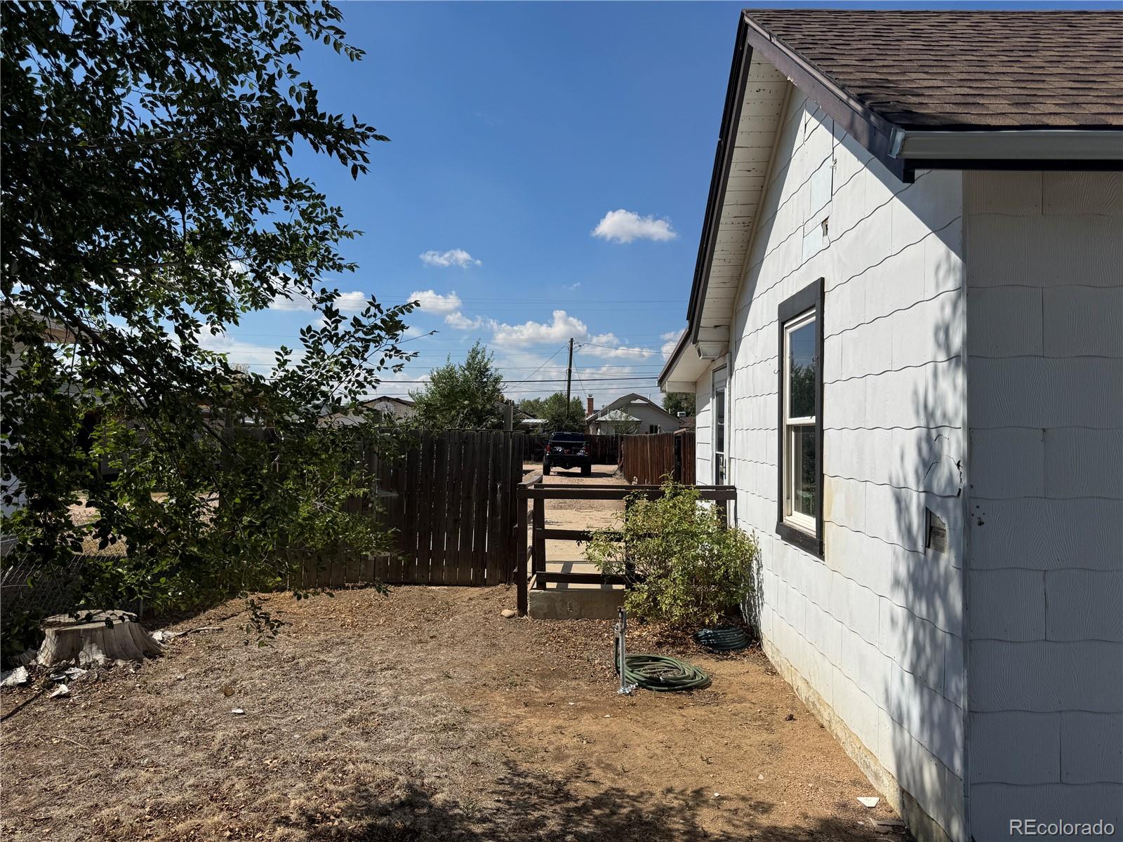 Side yard beside a white-sided home, with a wooden gate, sparse ground cover, and coiled hose.